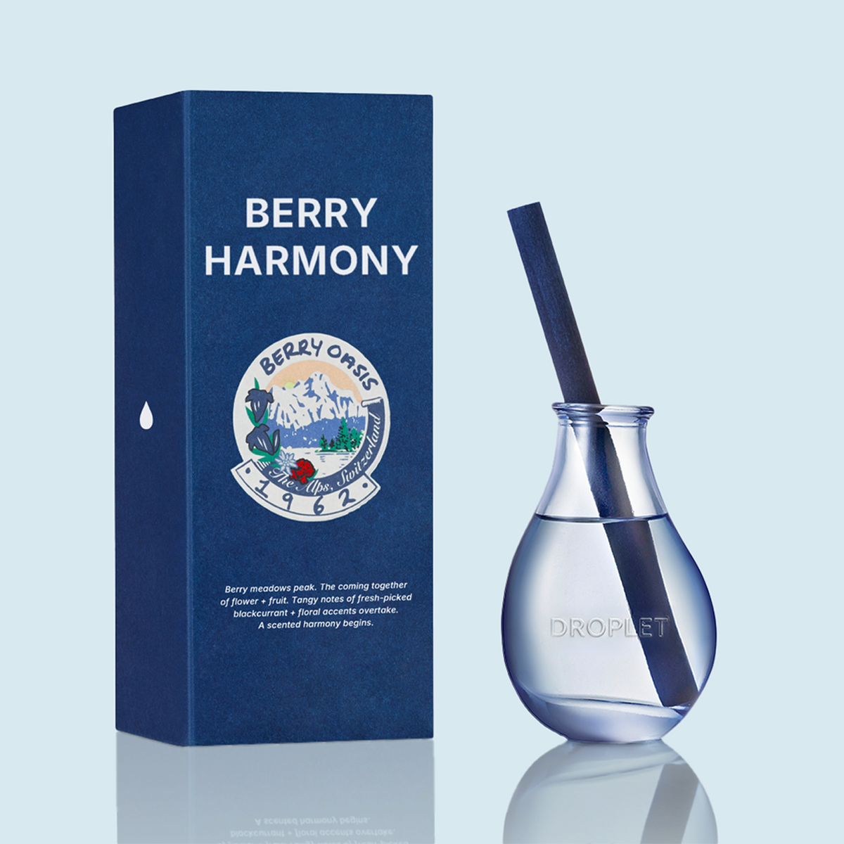 A Berry Harmony scented reed diffuser bottle with a dark blue reed and clear glass body, shown next to its dark blue packaging box on a light blue background.