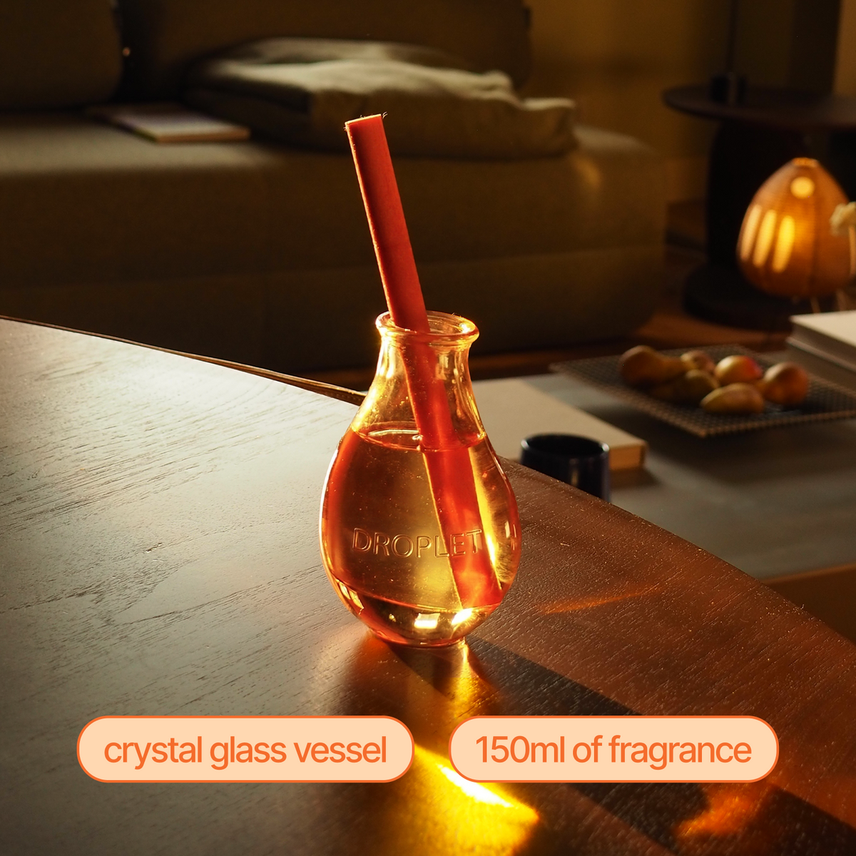 A small crystal glass vessel filled with amber-colored fragrance, with a red cylindrical straw inserted into it, placed on a wooden table in a warmly lit room. Text labels at the bottom read 'crystal glass vessel' and '150ml of fragrance'.