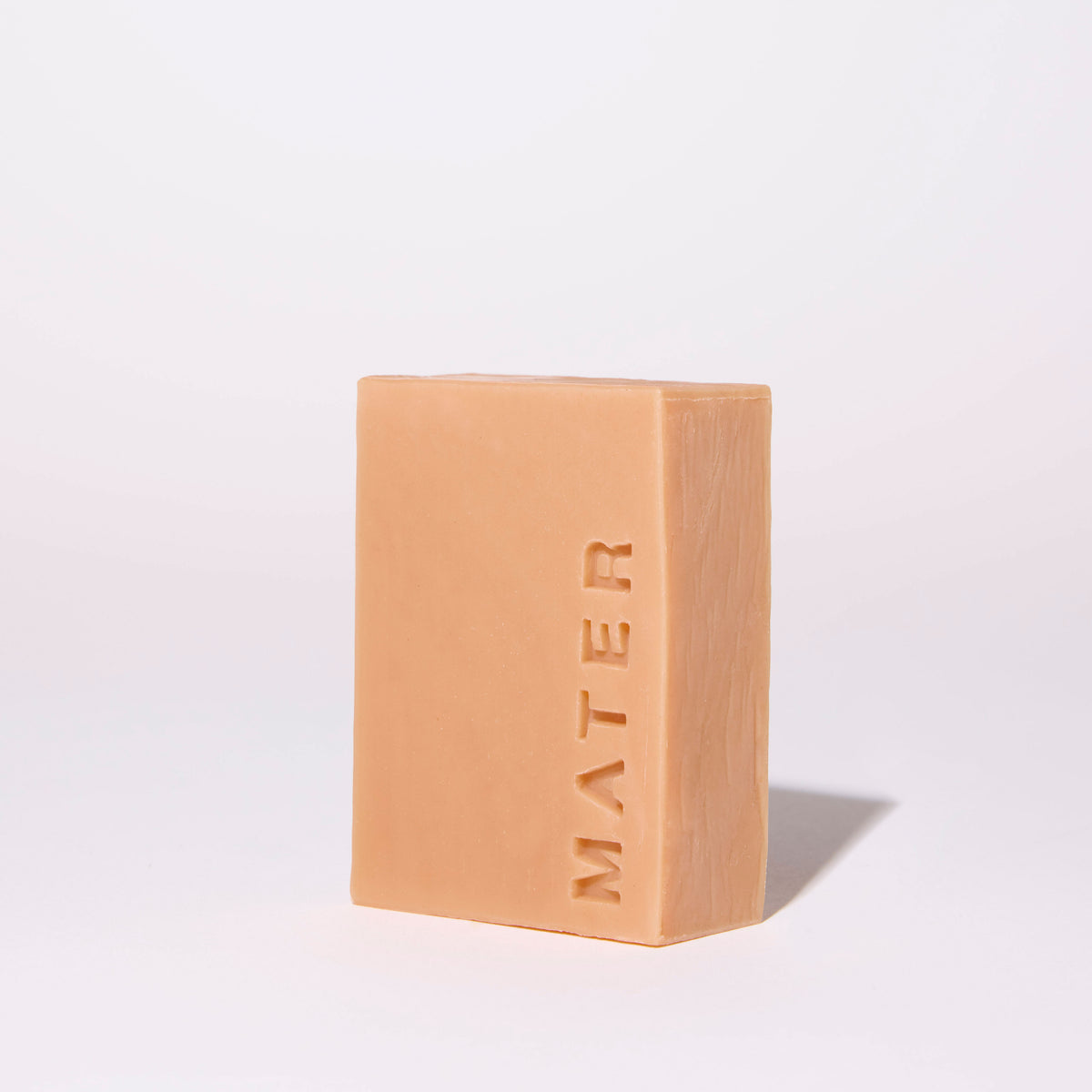 A rectangular bar of MATER soap in a warm peach-beige color, standing upright on a white studio background with a soft shadow cast to the right.