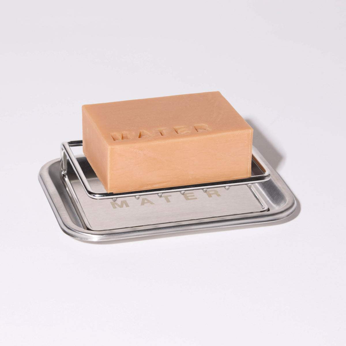 A rectangular bar of light peach-colored soap resting on a square stainless steel soap dish, photographed on a clean white background.