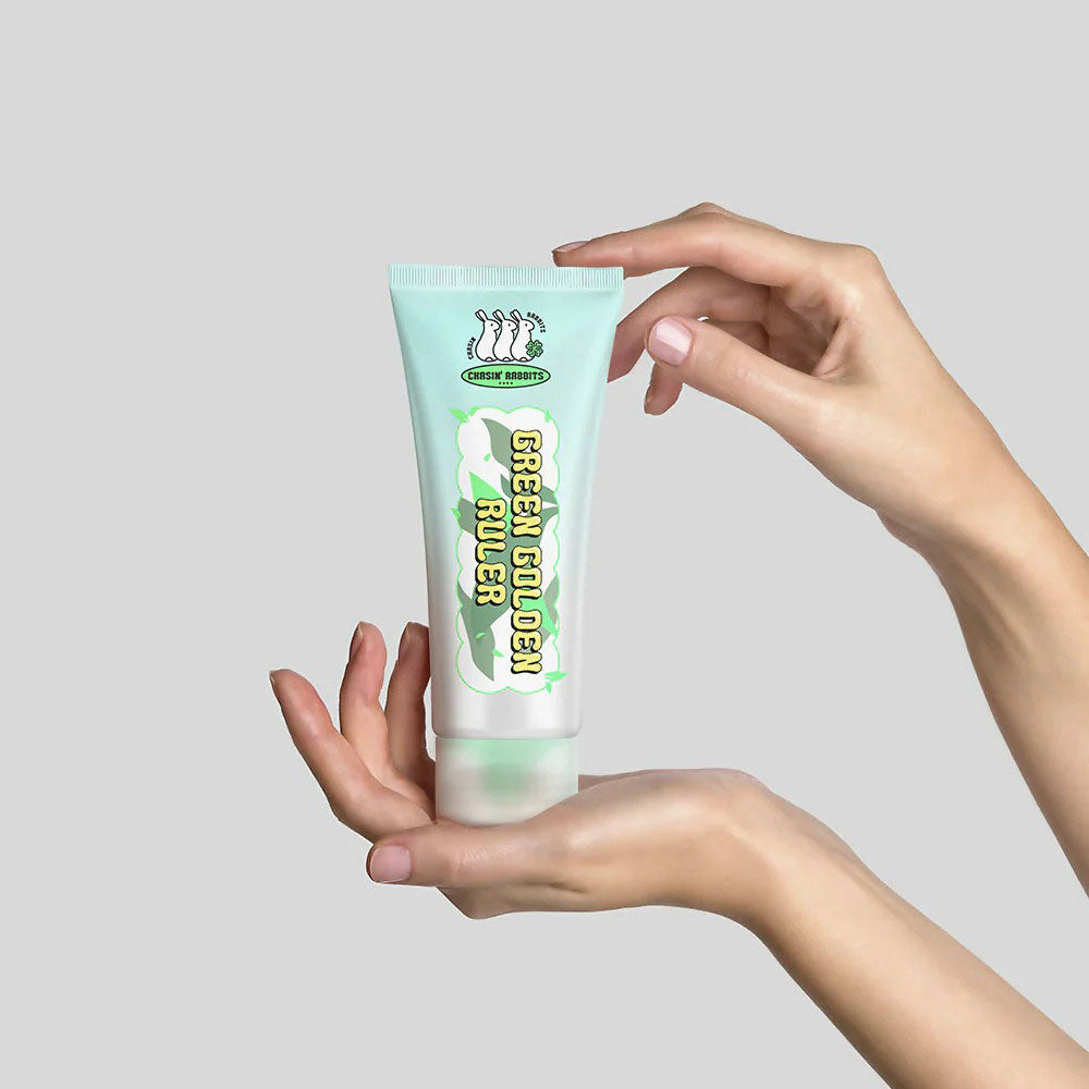 Hand holding a tube of 'Green Golden Rule' cream against a gray background