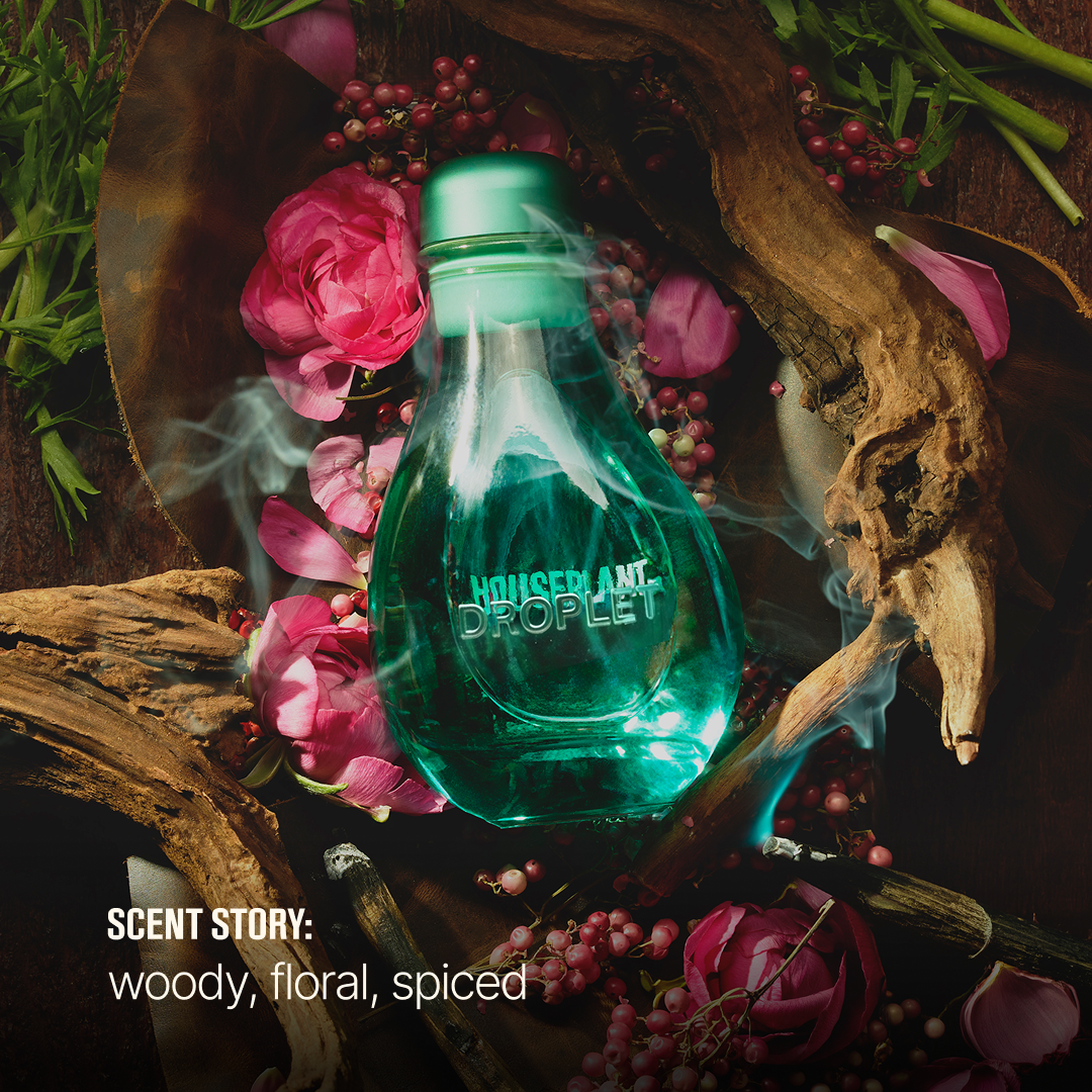 A clear glass bottle of Houseplant Droplet fragrance with a translucent teal-green cap and body, surrounded by pink roses, red berries, green leaves, smoke, and dark wood props. Text at the bottom reads 'SCENT STORY: woody, floral, spiced'.