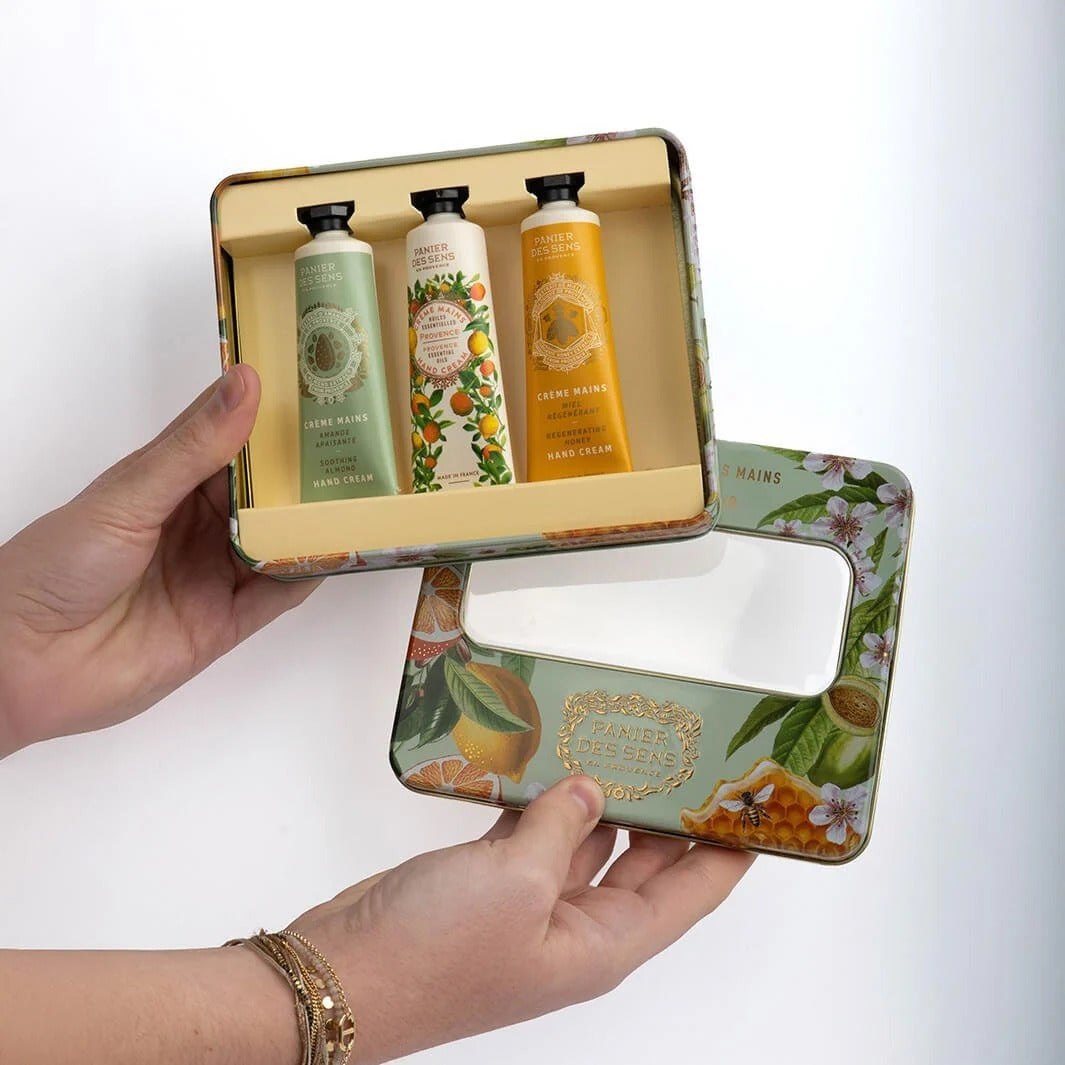 Hand holding a decorative tin with three hand creams inside on a white background.
