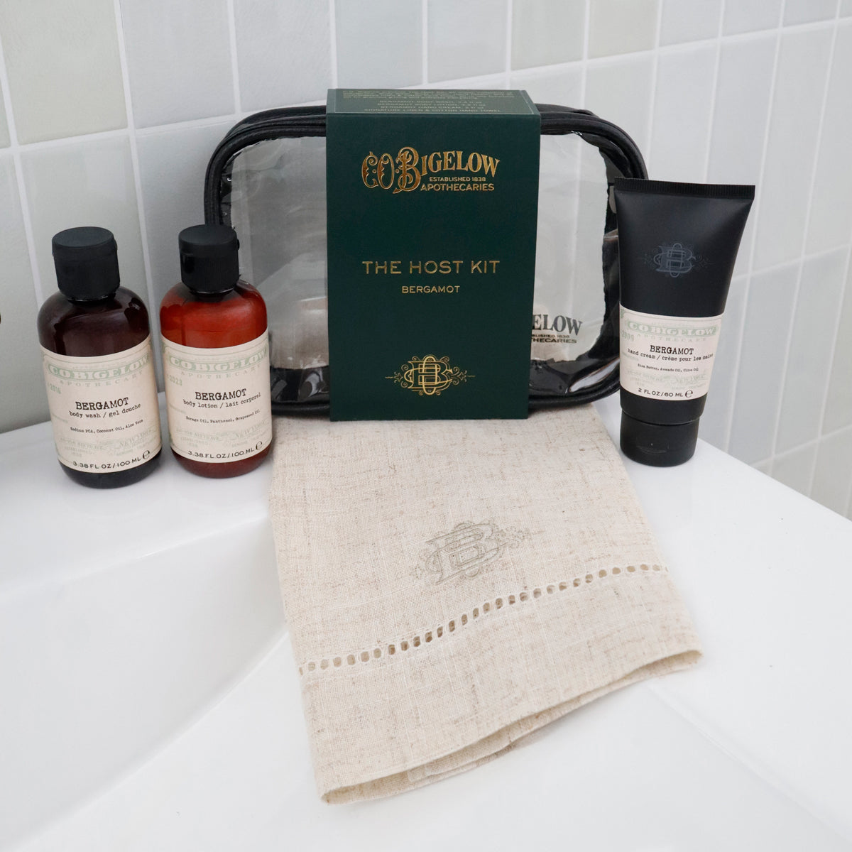 Bigelow Apothecary 'The Host Kit' Bermuda travel set arranged on a white bathroom counter, including two amber bottles, a black tube, a dark green box, a black zip pouch, and a beige linen towel.