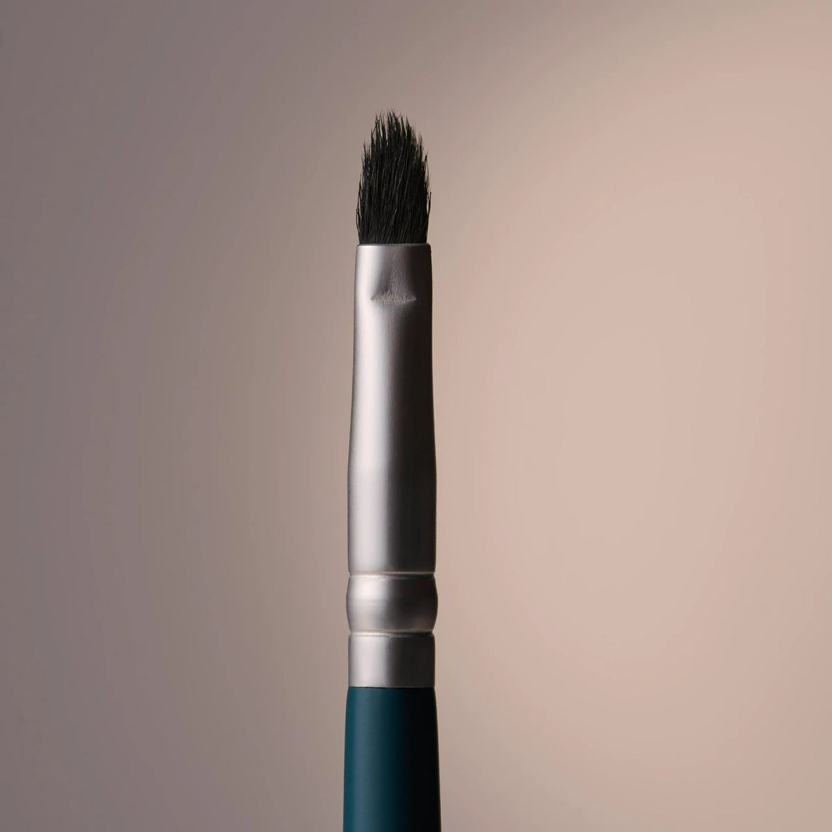 A close-up studio shot of makeup brush with dark bristles, a silver ferrule, and a dark teal handle against a soft beige gradient background.