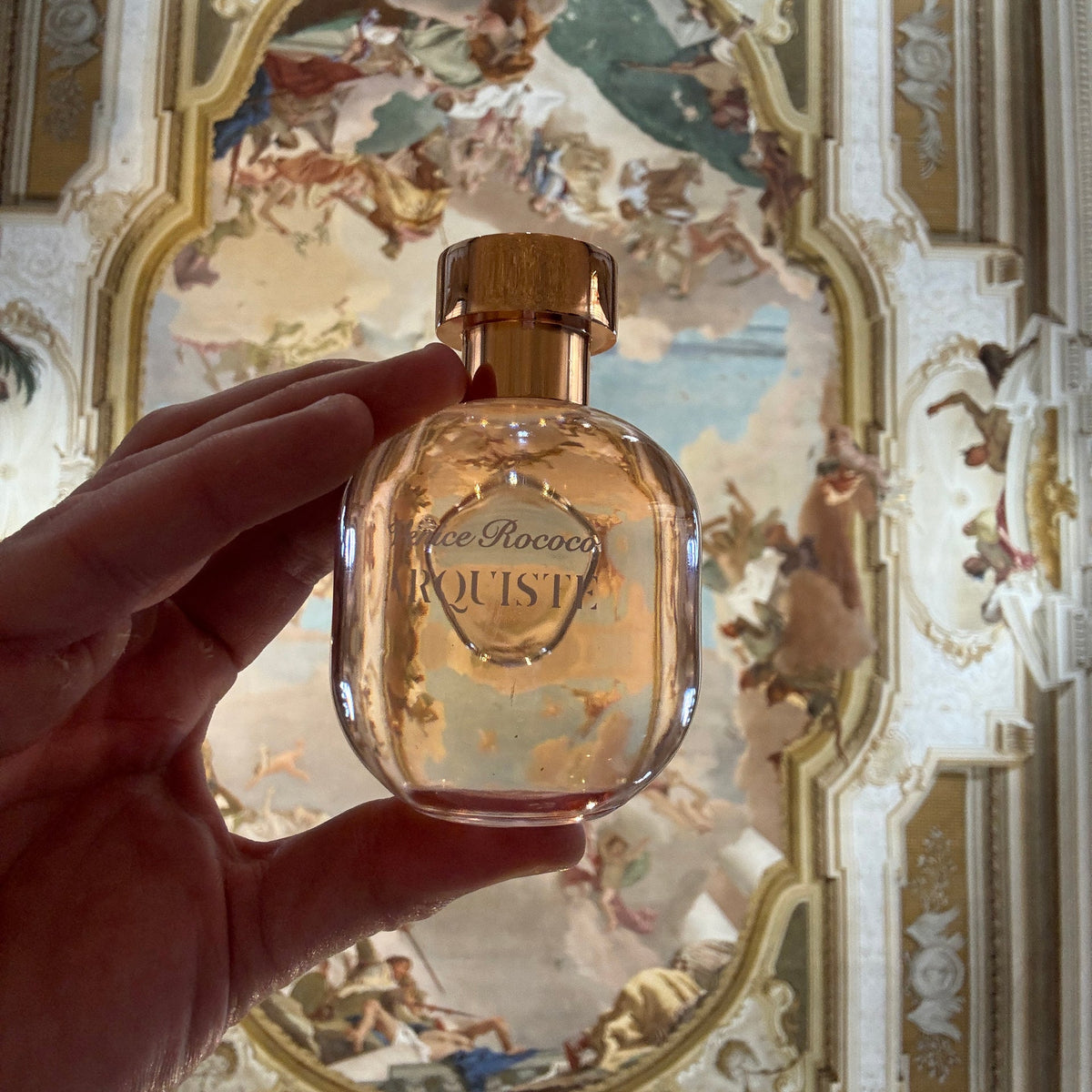 A hand holding a bottle of Arquiste Venice Rococo perfume in front of a painting