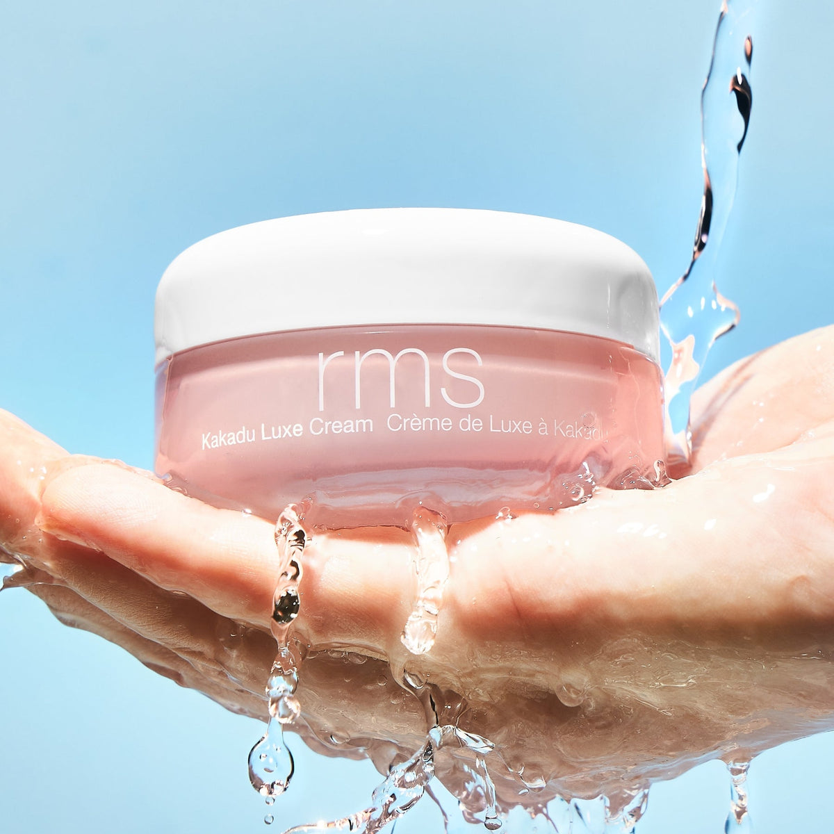Hand holding RMS Kakadu Luxe Cream jar that is being splashed by water.