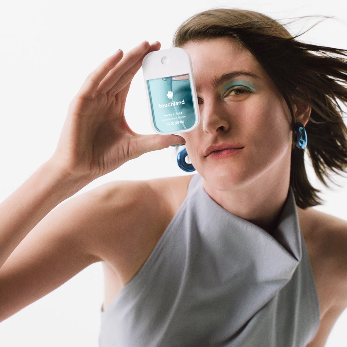 A woman in a light gray halter top holds Touchland Power Mist Hydrating Hand Sanitizer - Blue Sandalwood, in front of her face, on a white background