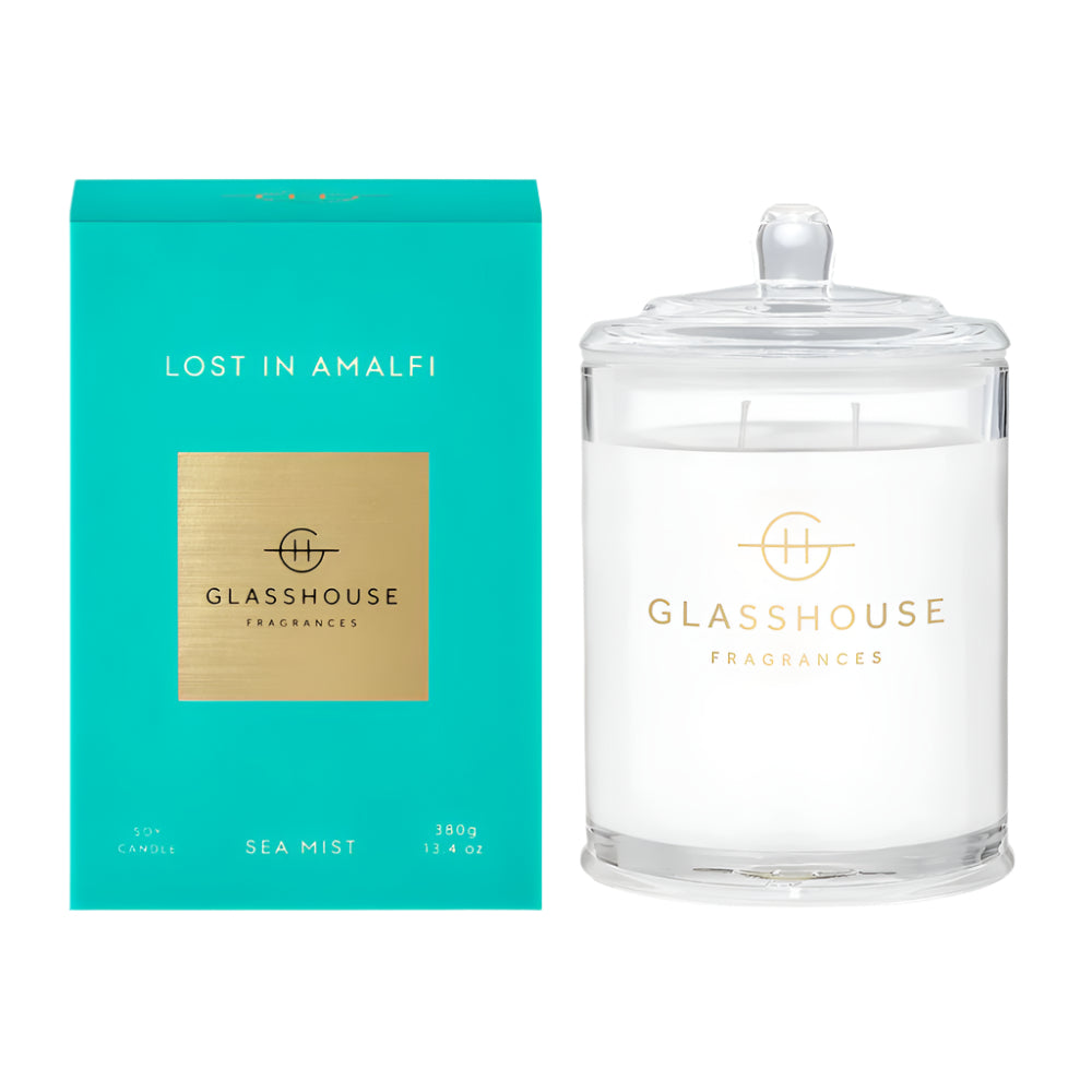Glasshouse Fragrances Lost in Amalfi Candle is shown with its lid on next to the light Aqua-blue box it comes in.  It is a clear glass vessel with a lid and Glasshouse Fragrances Logo on the vessel. 
