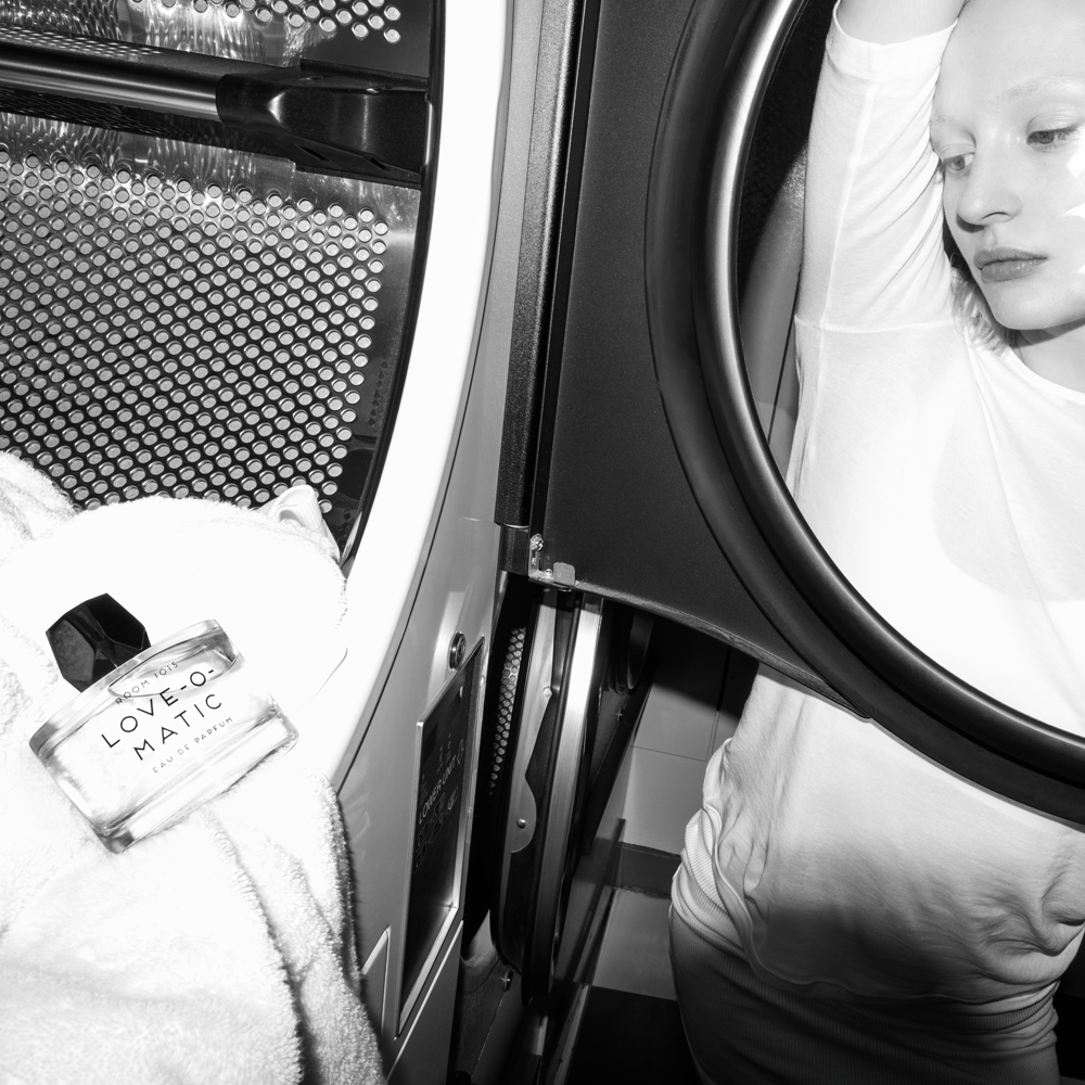 Woman in white looks through washing machine glass window at bottle of Room 1015 Love-O-Matic Eau de Parfum resting on a white towel.