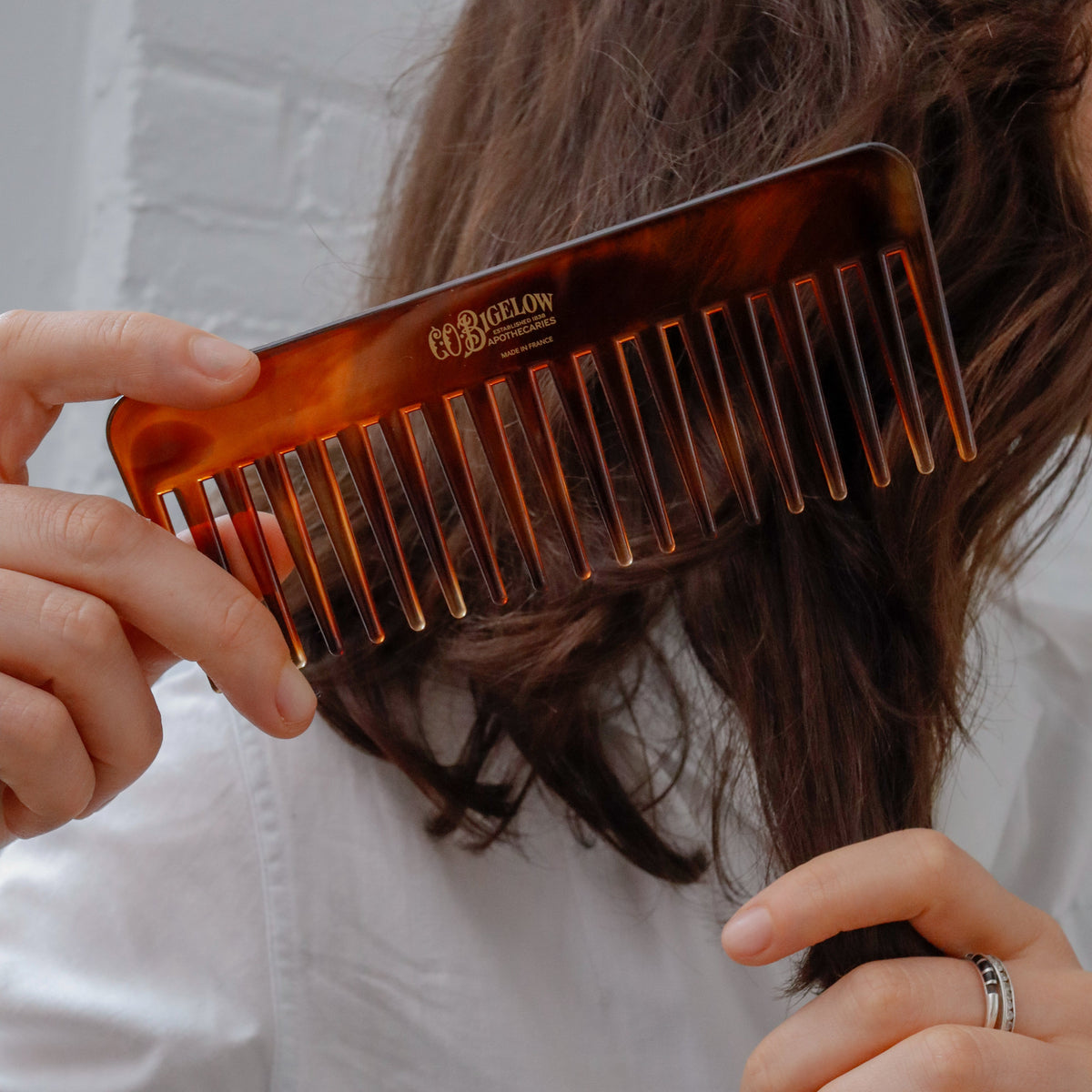 A close-up of a person holding a wide-tooth brown tortoiseshell hair comb over their dark brown hair, wearing a white shirt against a white brick wall background.