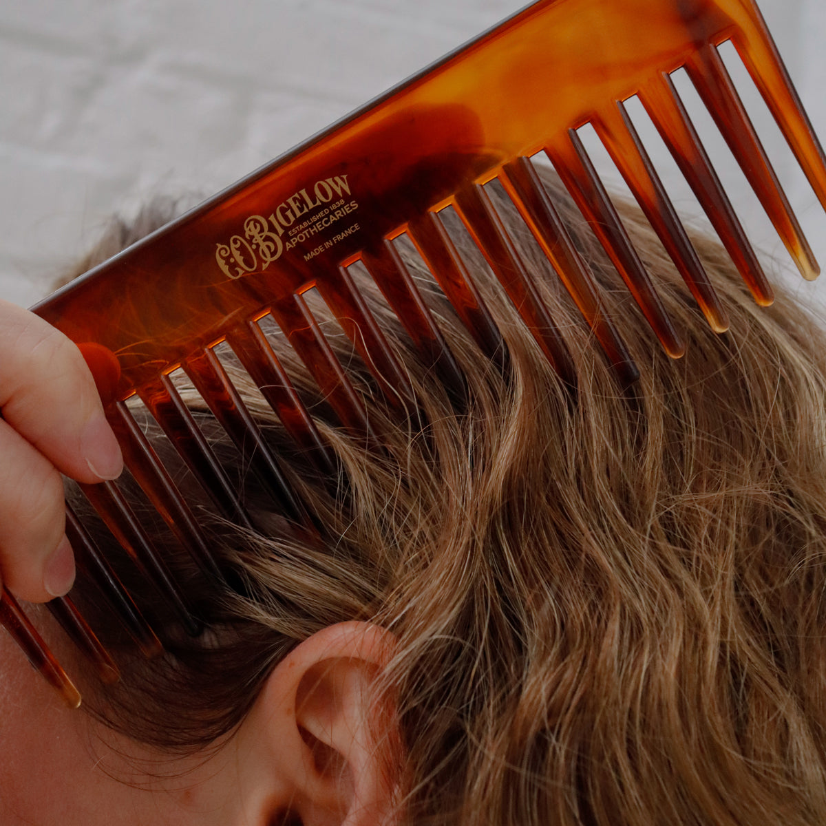 A close-up of a hand holding a translucent amber-brown hair comb against a person's light brown hair. The brand name 'ÉBICELON' is printed in gold on the comb.