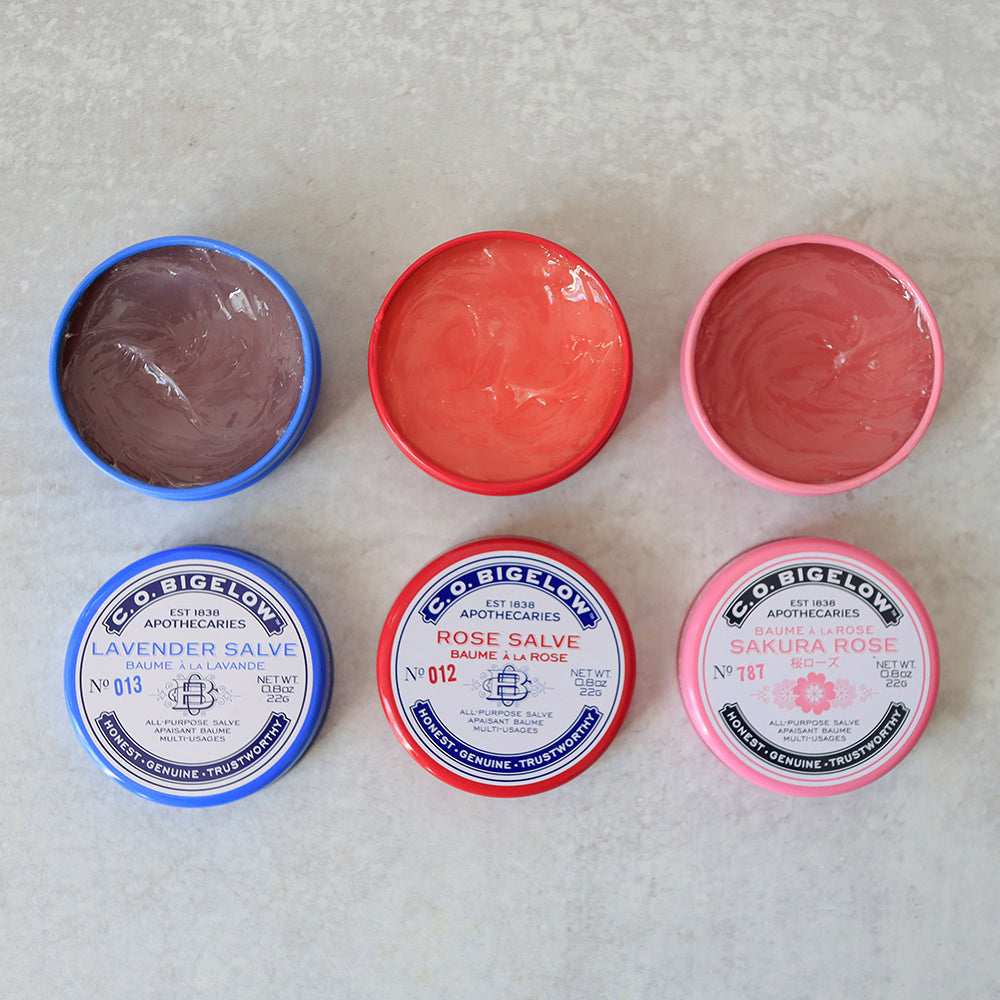 Six containers of C.O. Bigelow salves in different colors on a textured surface.
