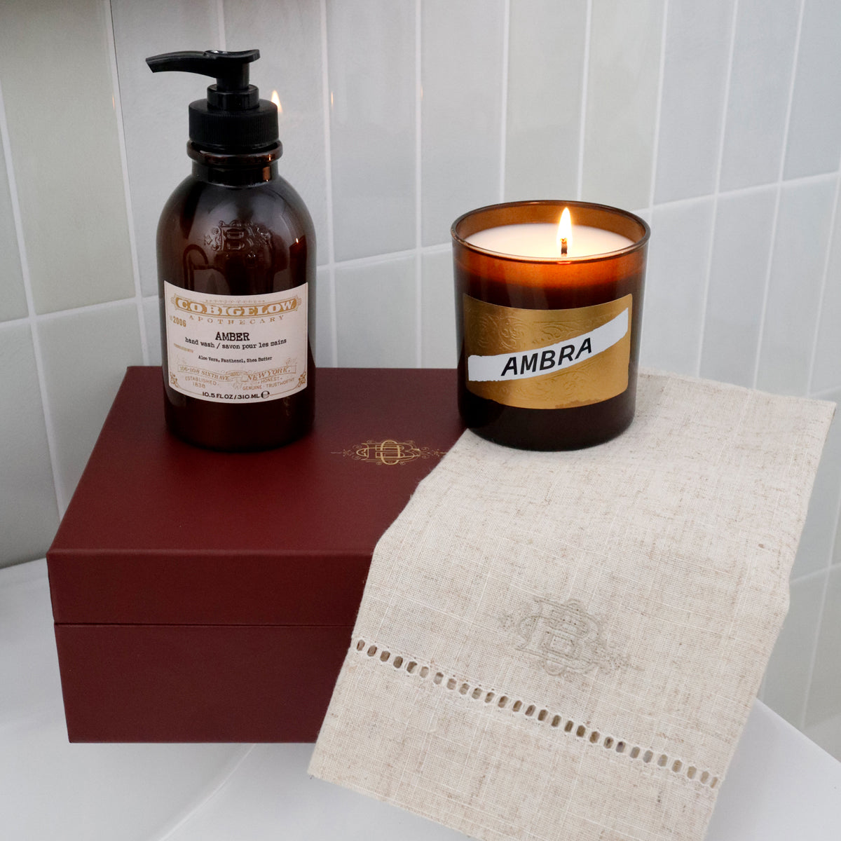 A candle and a pump bottle from the brand CO Bigelow, both labeled 'Ambra', arranged on a white surface with a dark red gift box and a beige linen cloth against a white tiled wall.
