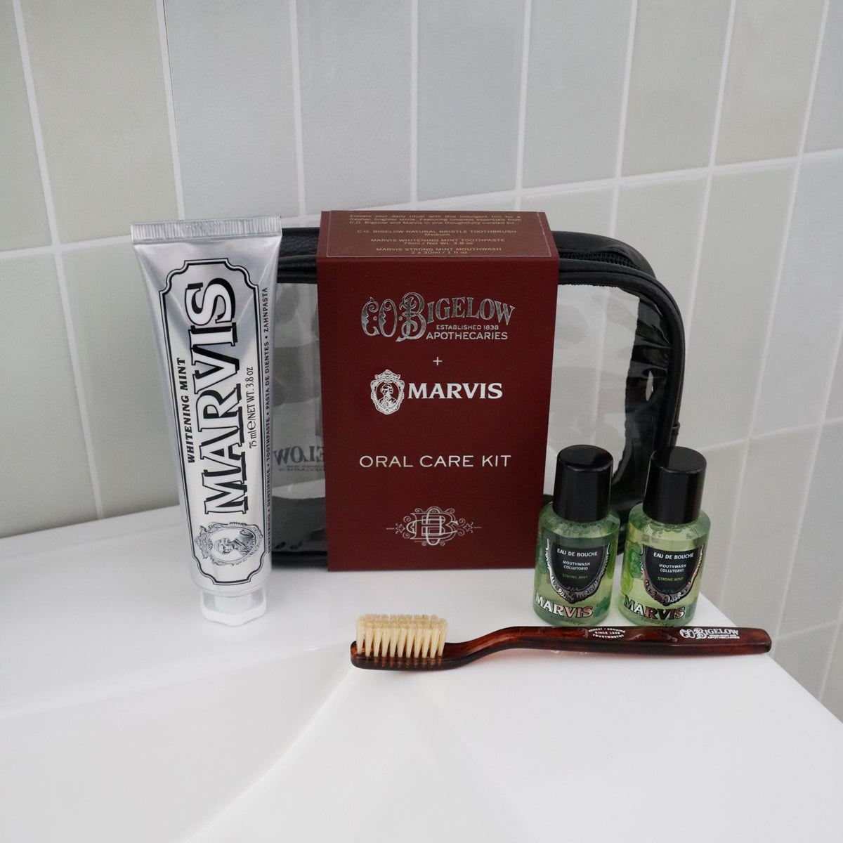 A Marvis oral care kit arranged on a white bathroom counter against a white tiled wall, including a silver tube of mint toothpaste, two small green liquid bottles, and a brown toothbrush.