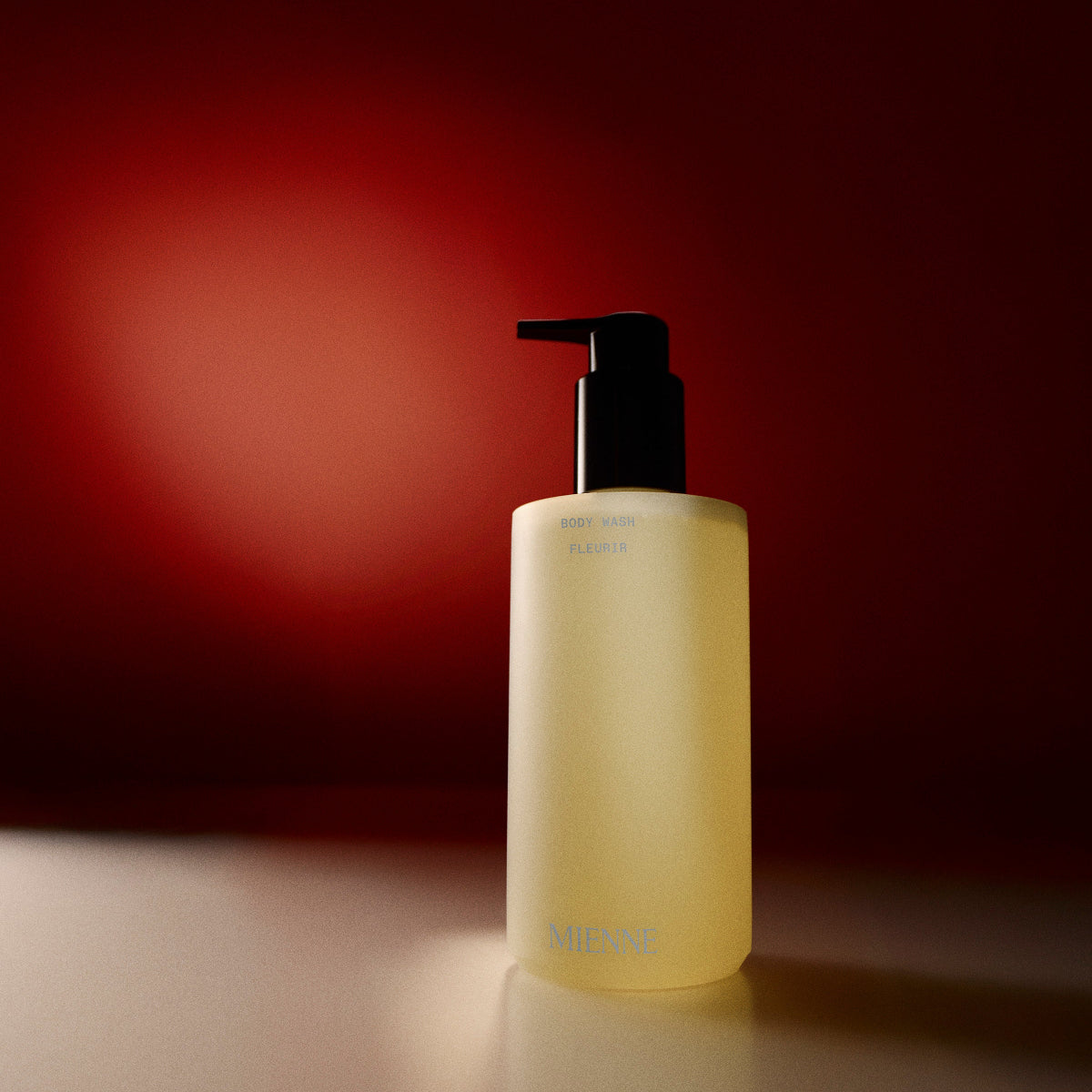 Mienne Body Wash bottle in the scent Fleurir on a tan surface in front of a red background.