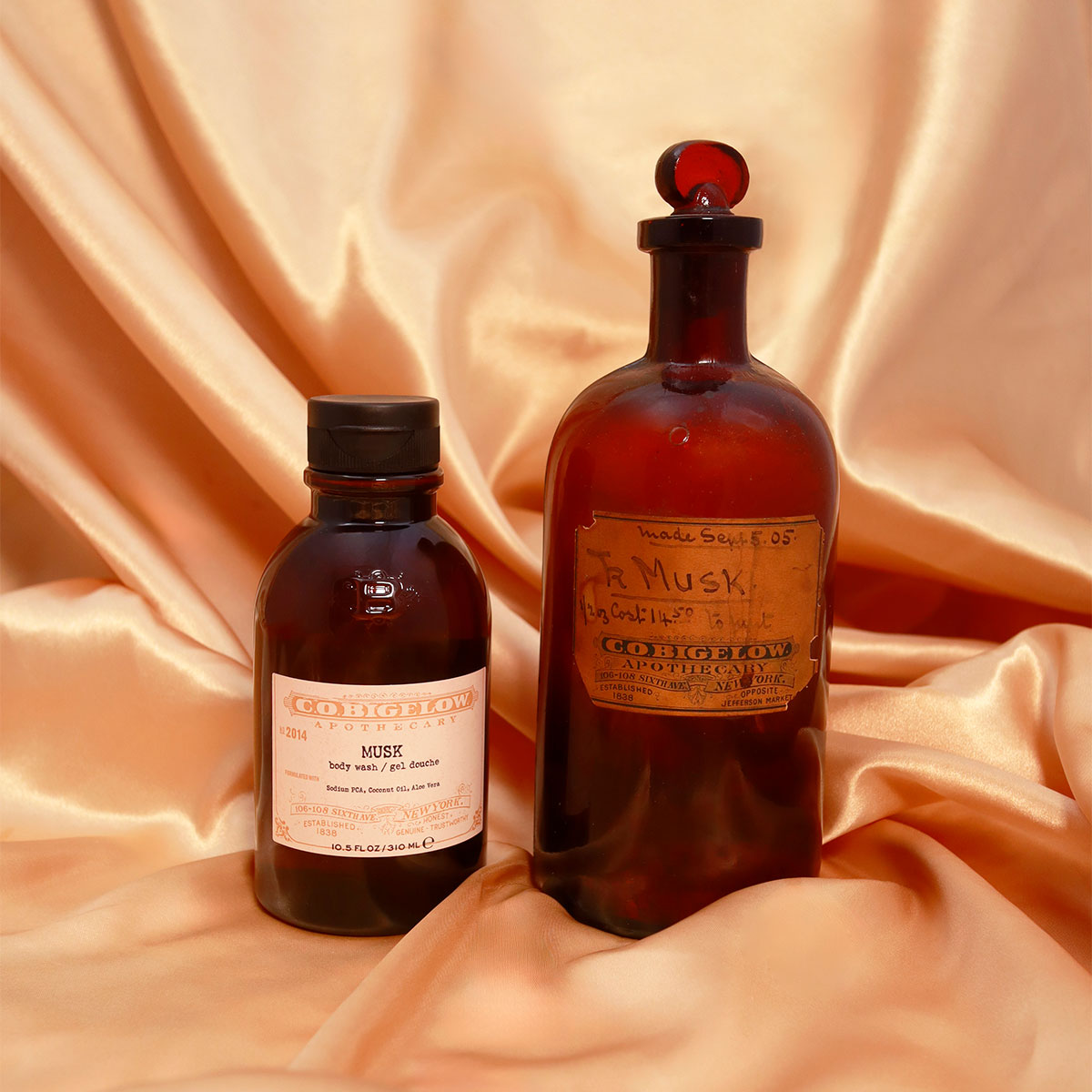 C.O. Bigelow Musk Body Wash in an Amber bottle next to an Original C.O. Bigelow Musk Apothecary Jar on top of draped silk.