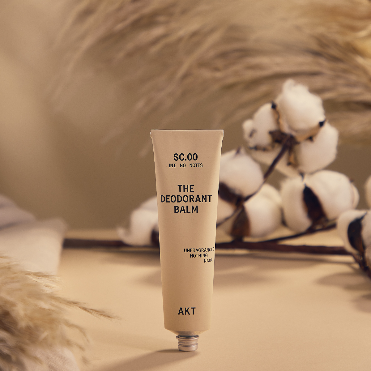 AKT unscented Deodorant Balm Tube standing on a tan surface in front of cotton plants and wheat. 