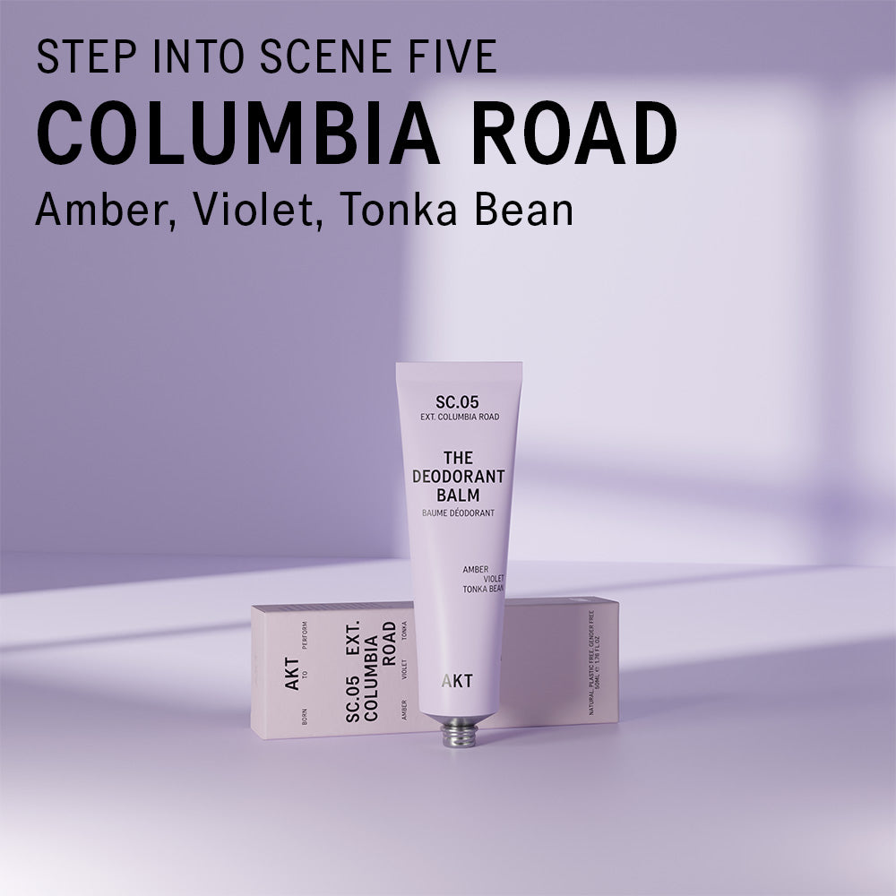 AKT Columbia Road scented deodorant balm standing on and in front of a lilac colored surface. Text overlaid on the image above the product reads "Step into scene five. Columbia Road. Amber, Violet, Tonka Bean."