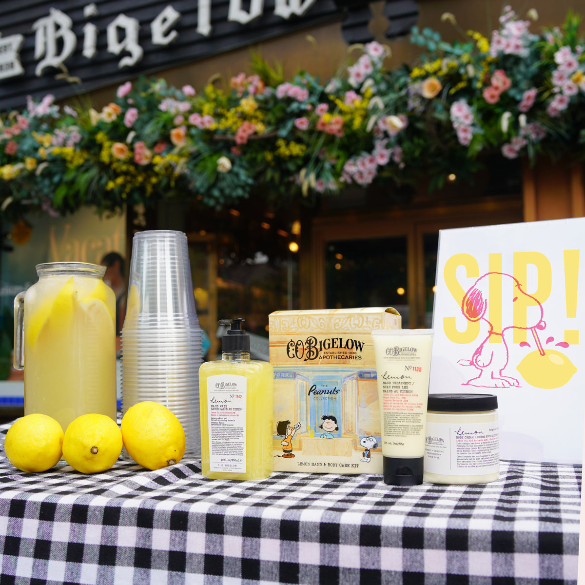 A table setup in front of a storefront with flower garlands, featuring a pitcher of lemonade, stacked clear cups, three lemons, and several C.O. Bigelow products including a hand wash, lotion, and jar on a black-and-white checkered tablecloth. A white sign with a yellow cartoon character and the word 'SIP!' is visible on the right.