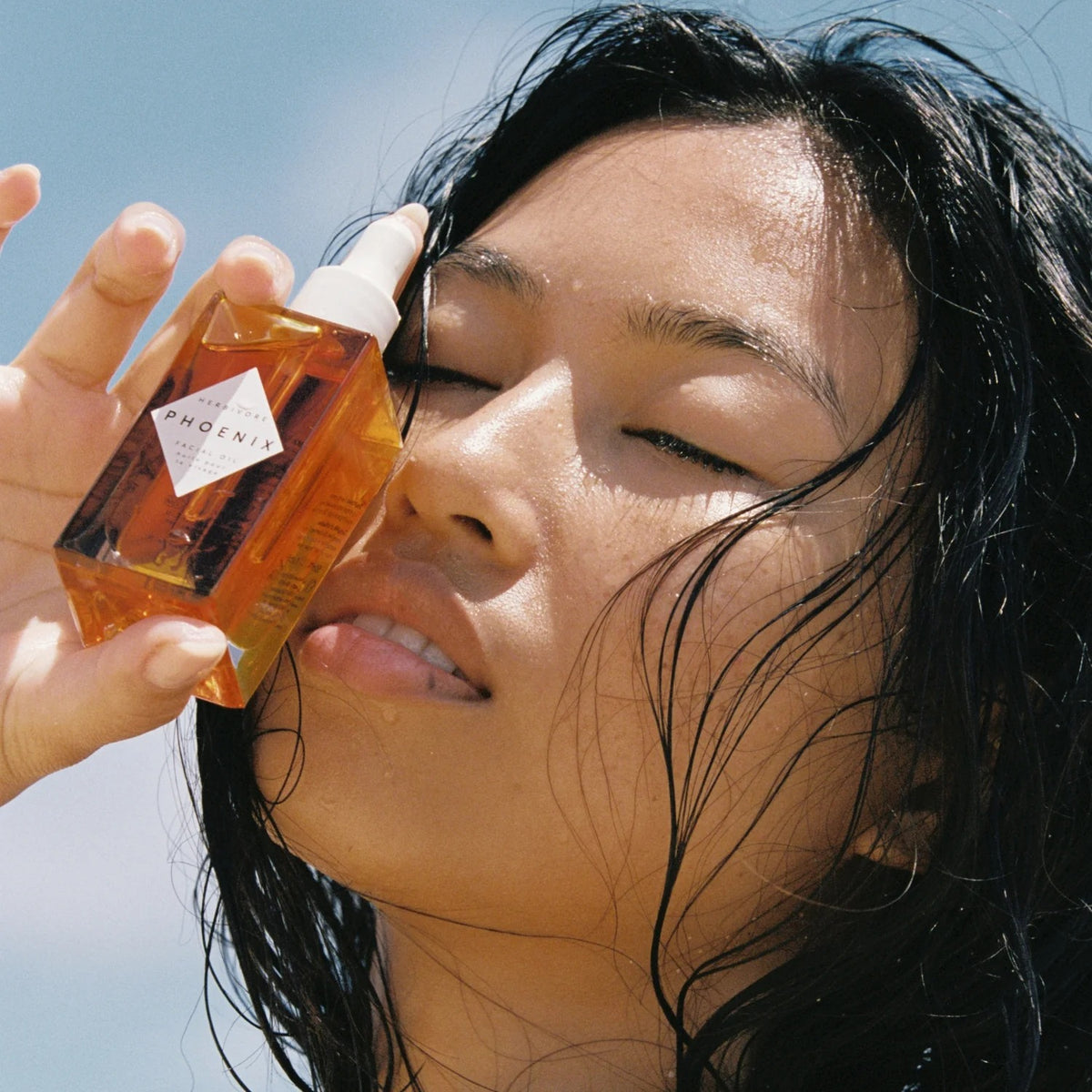 Person holding a bottle of 'Phoenix' skincare product near their face with a clear blue sky background