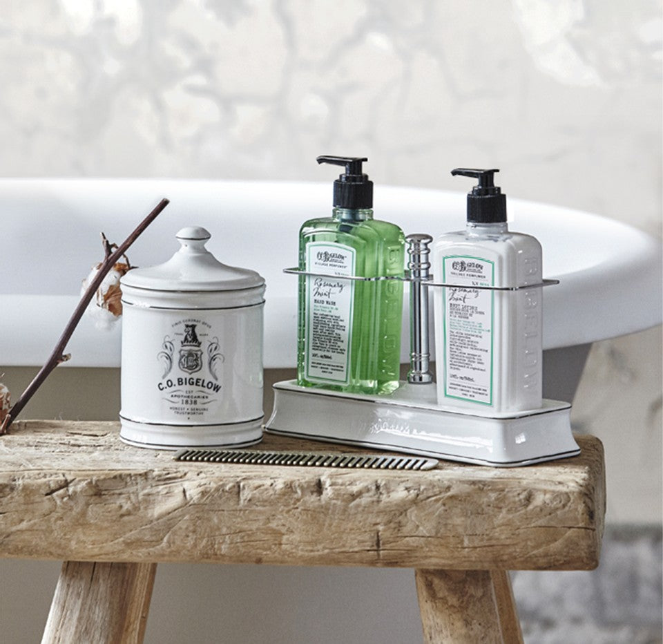 A rustic wooden stool holding a white ceramic apothecary jar labeled 'C.O. Bigelow, a tray with CO Bigelow Rosemary Mint Hand Wash and Body Lotion pump bottles. A freestanding bathtub is blurred in the background.