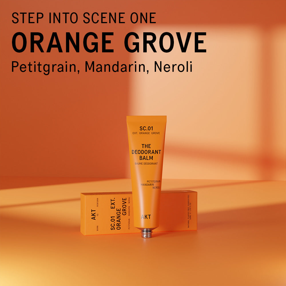 AKT Orange Grove Deodorant Balm standing on and in front of an orange surface. Text overlaid on the image above the product reads "Step into scene one. Orange Grove. Petitgrain, Mandarin, Neroli."