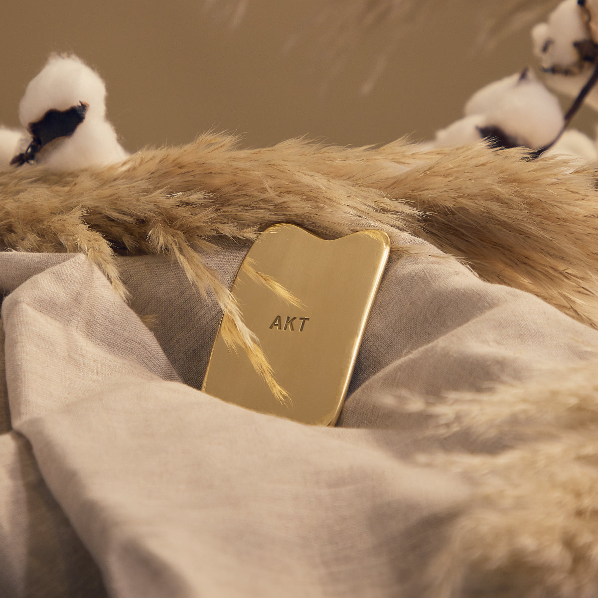 AKT's Gua Sha Applicator laying on tan linen in front of wheat and cotton plants. 