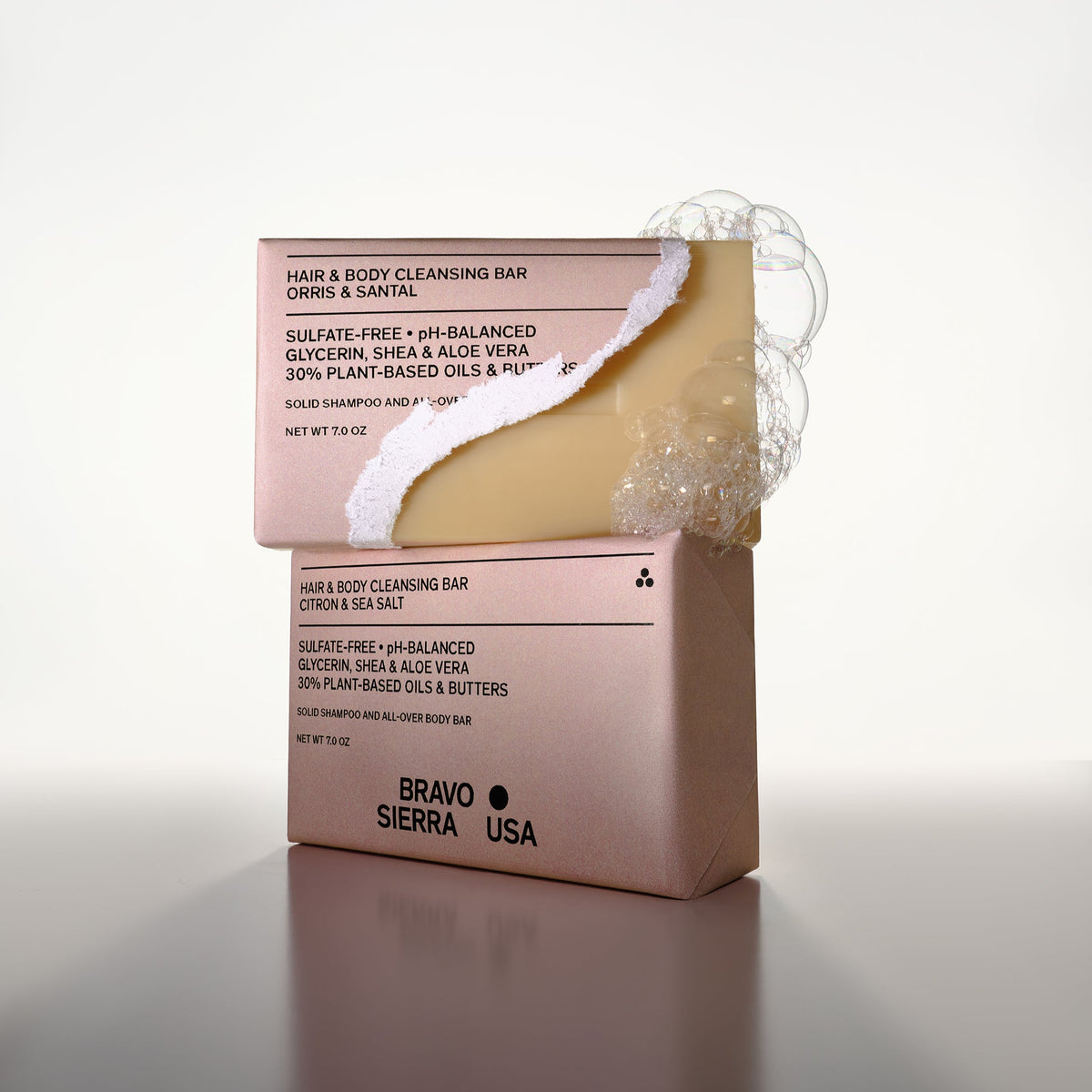 Bravo Sierra Orris & Santal Cleansing Bar Soap sitting atop of the Bravo Sierra Citron & Sea Salt Cleansing Bar Soap. The Orris & Santal bar packaging is ripped half open revealing the naked bar and soapy bubbles.