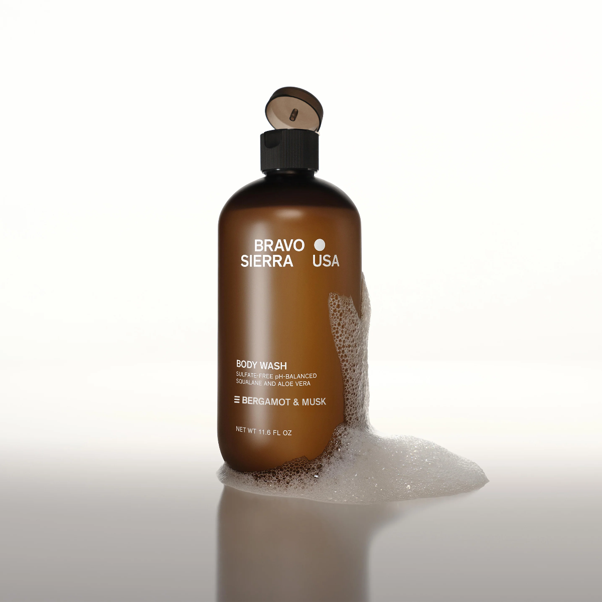 Bravo Sierra Bergamot & Musk Body Wash next to soap suds sitting on a reflective surface in front of a white background.