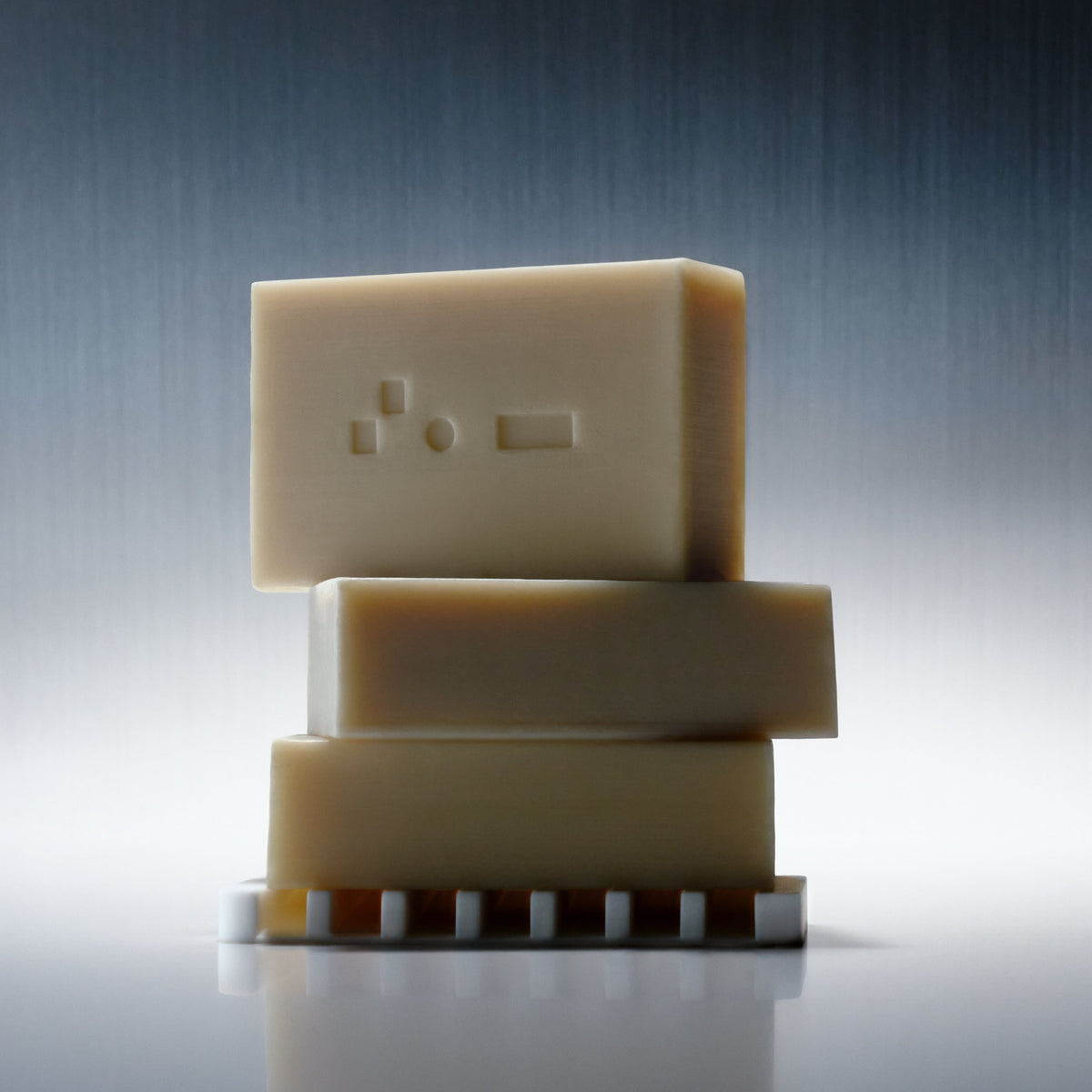 Three Bars of Bravo Sierra's Cleansing Bar Soaps stacked on top of one another on a soap dish in front of a white/gray background.