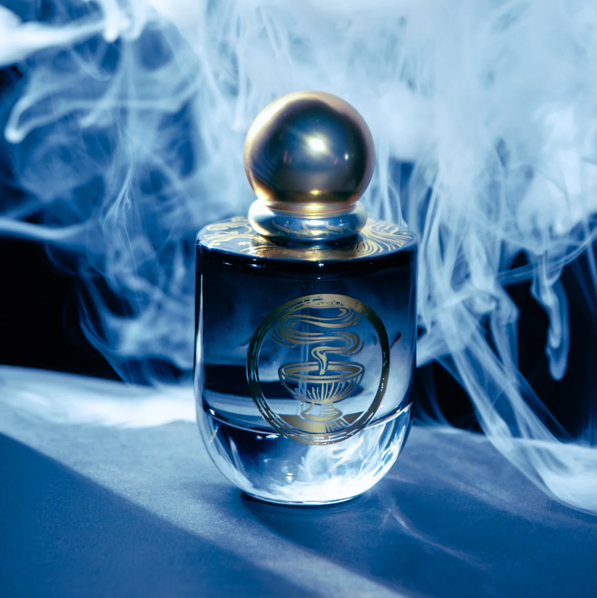 A clear glass perfume bottle with a metallic gold spherical cap and gold circular emblem on the front, set against a dark background with dramatic white smoke swirling around it.
