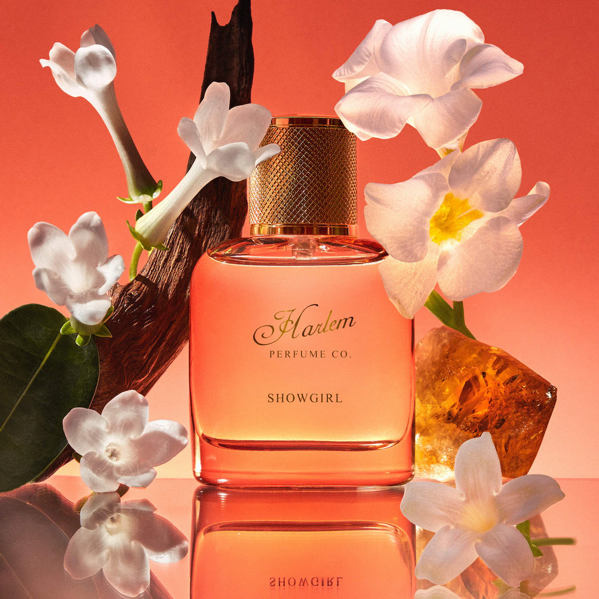 Perfume bottle labeled 'Harlem Perfume Co. Showgirl' surrounded by flowers on a reflective surface with a red-orange background.
