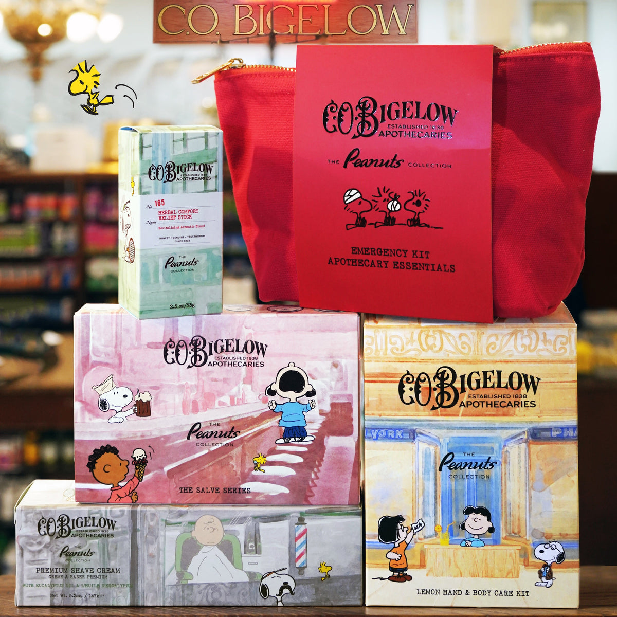 A coBigelow Apothecary Peanuts emergency kit display featuring multiple themed boxes and a red pouch on a store shelf. The collection includes an Emergency Kit box, a First Aid box, a Beauty Box, and a Hair & Body Care box, all illustrated with Peanuts characters, alongside a red drawstring pouch.