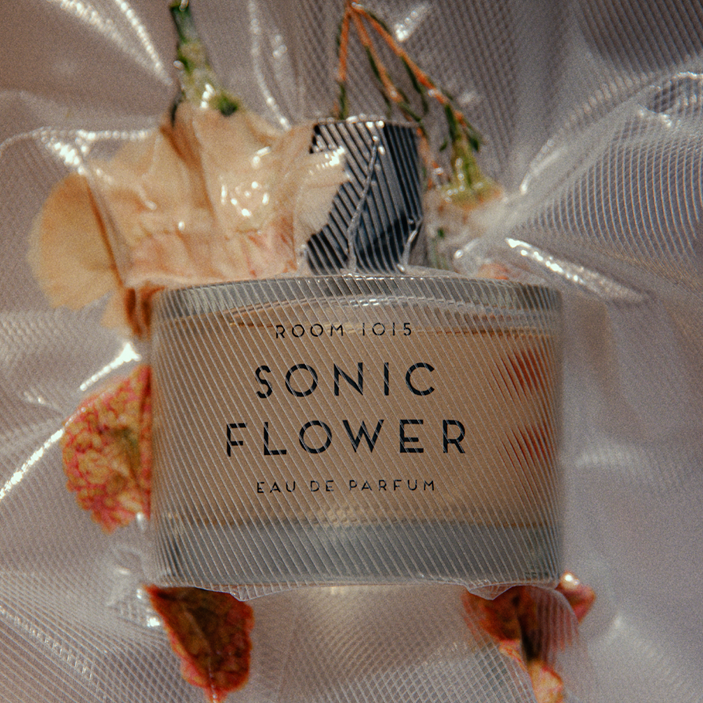 A perfume bottle labeled 'ROOM 1015 SONIC FLOWER EAU DE PARFUM' wrapped in a translucent mesh bag with dried flowers around it, on a light neutral background.