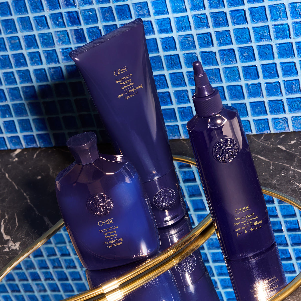 Oribe Supershine Shampoo and Conditioner and Mirror Rinse Glass Hair Treatment on mirrored surface in front of a blue tile backsplash.
