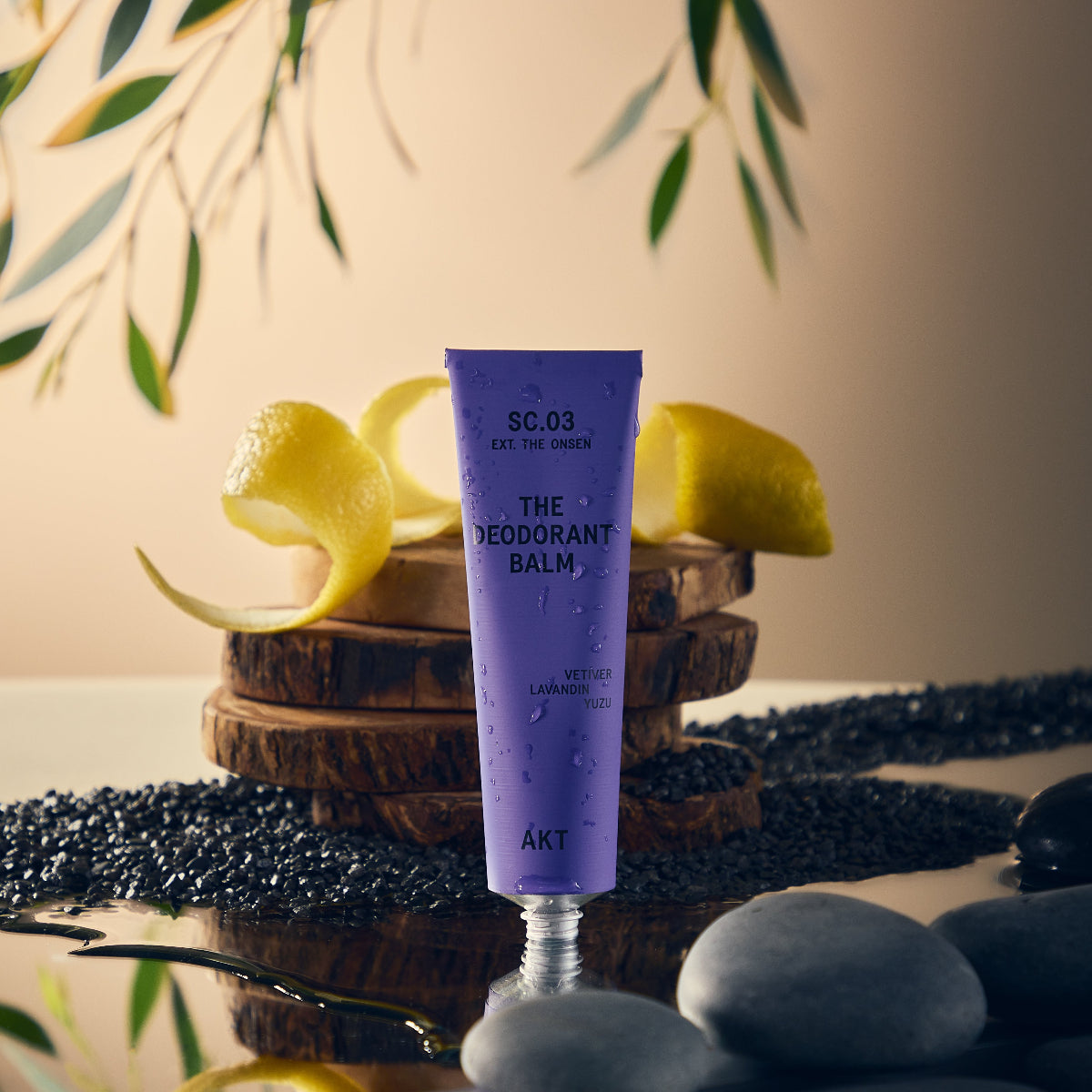 AKT The Onsen Scented Deodorant Balm Tube standing on a mirrored surface surrounded by wood, yuzu rinds, rocks, and green leaves. 