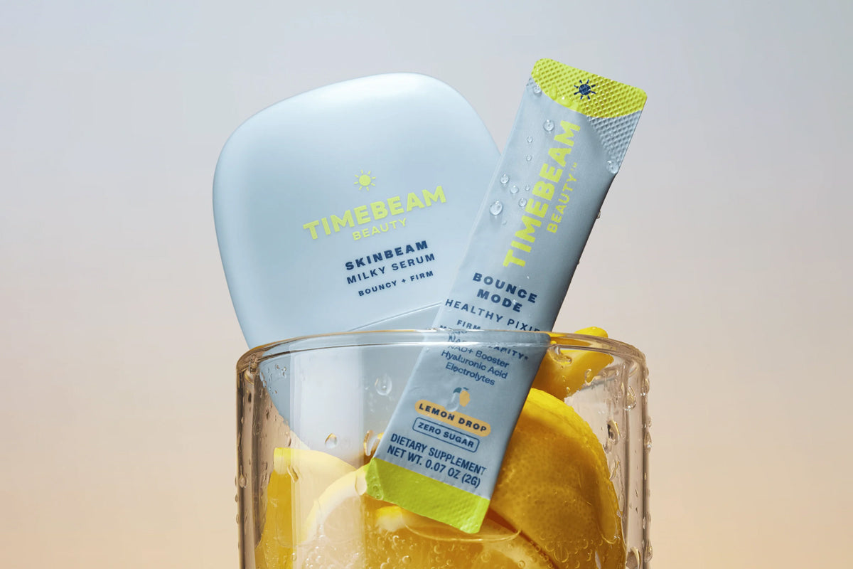 Timebeam skincare products displayed in a clear glass filled with ice and lemon slices: a large pale blue serum bottle and a slim blue packet with yellow-green accents labeled Bounce Mode Healthy Pixie.