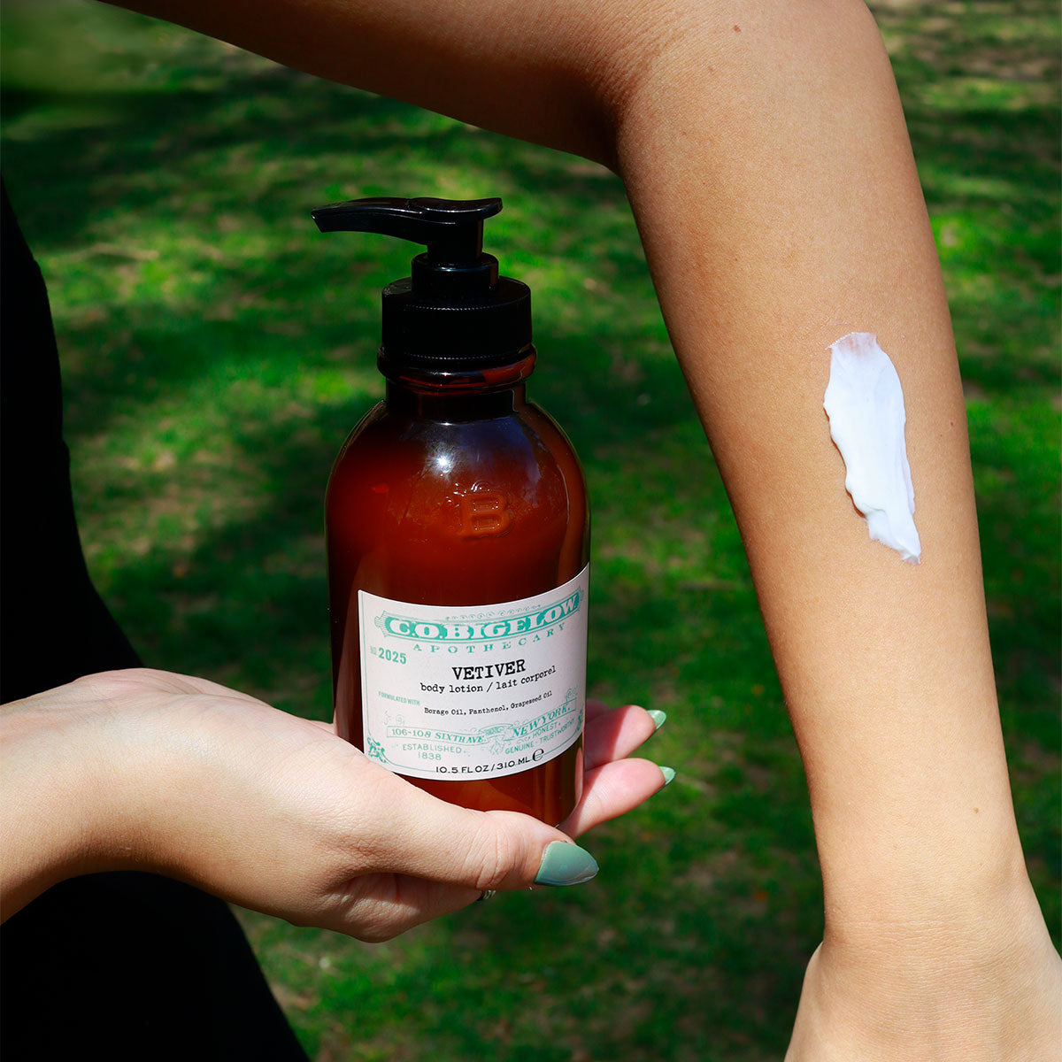 C.O. Bigelow Iconic Collection Vetiver Body Lotion bottle held in person's hand and swatched on their arm in front of green grass. 