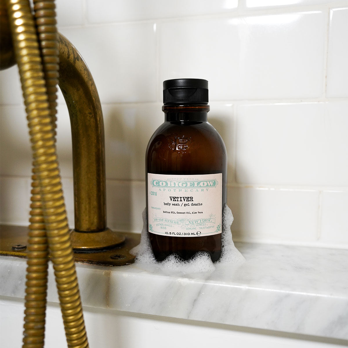 C.O. Bigelow Iconic Collection Vetiver Body Wash bottle on a bathtub ledge dripping with soap suds. 