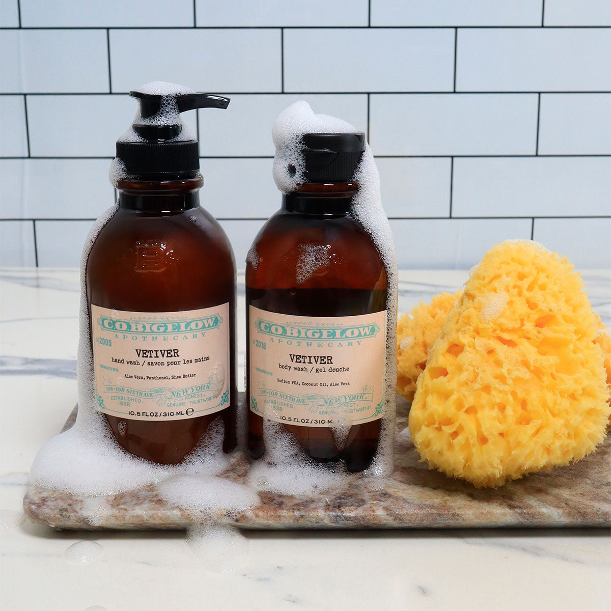 C.O. Bigelow Vetiver Hand Wash and Body Wash on a granite tray next to soapy sponges atop a bathroom counter. 