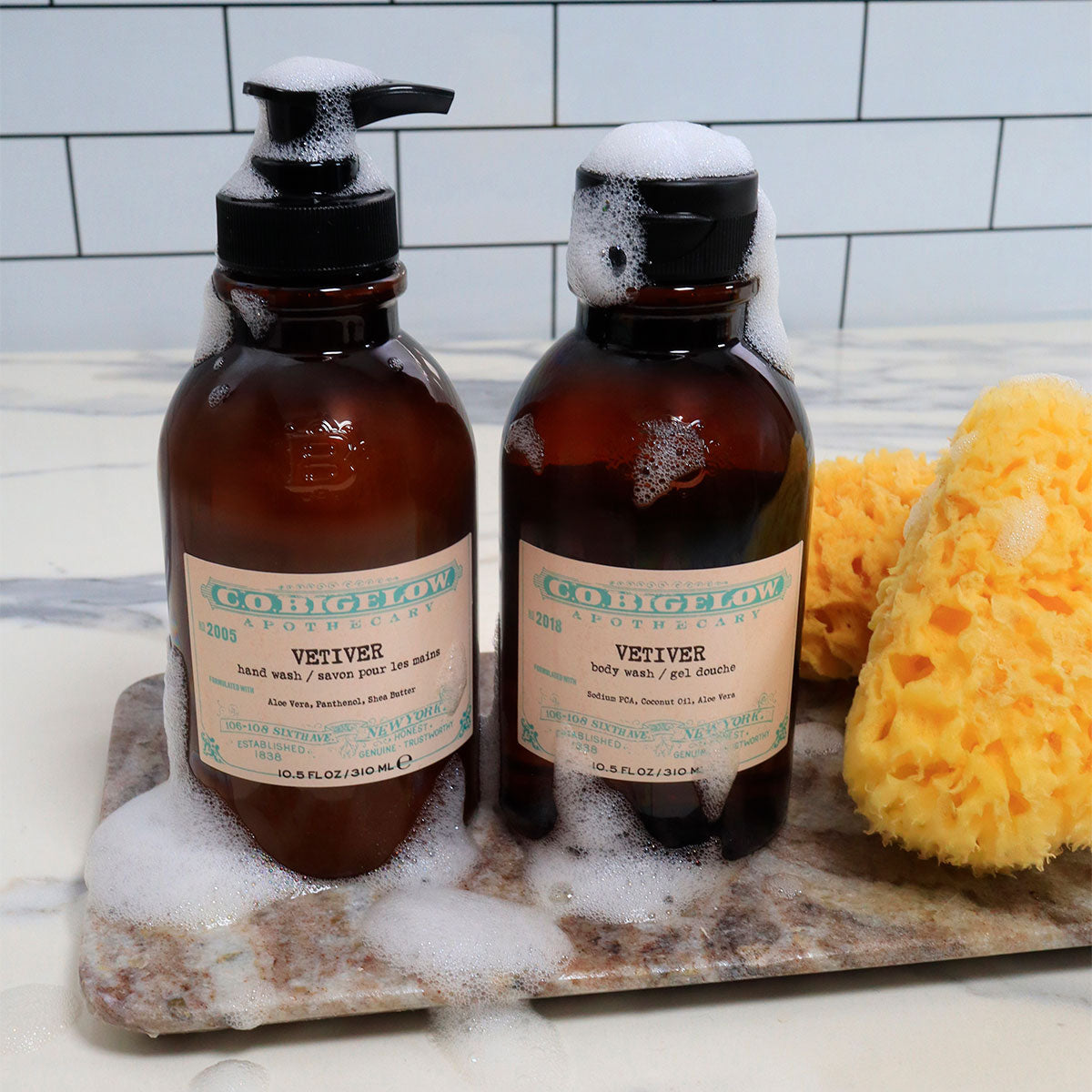 C.O. Bigelow Vetiver Hand Wash and Vetiver Body Wash on top of a granite tray with soapy sponges on a bathroom countertop. 