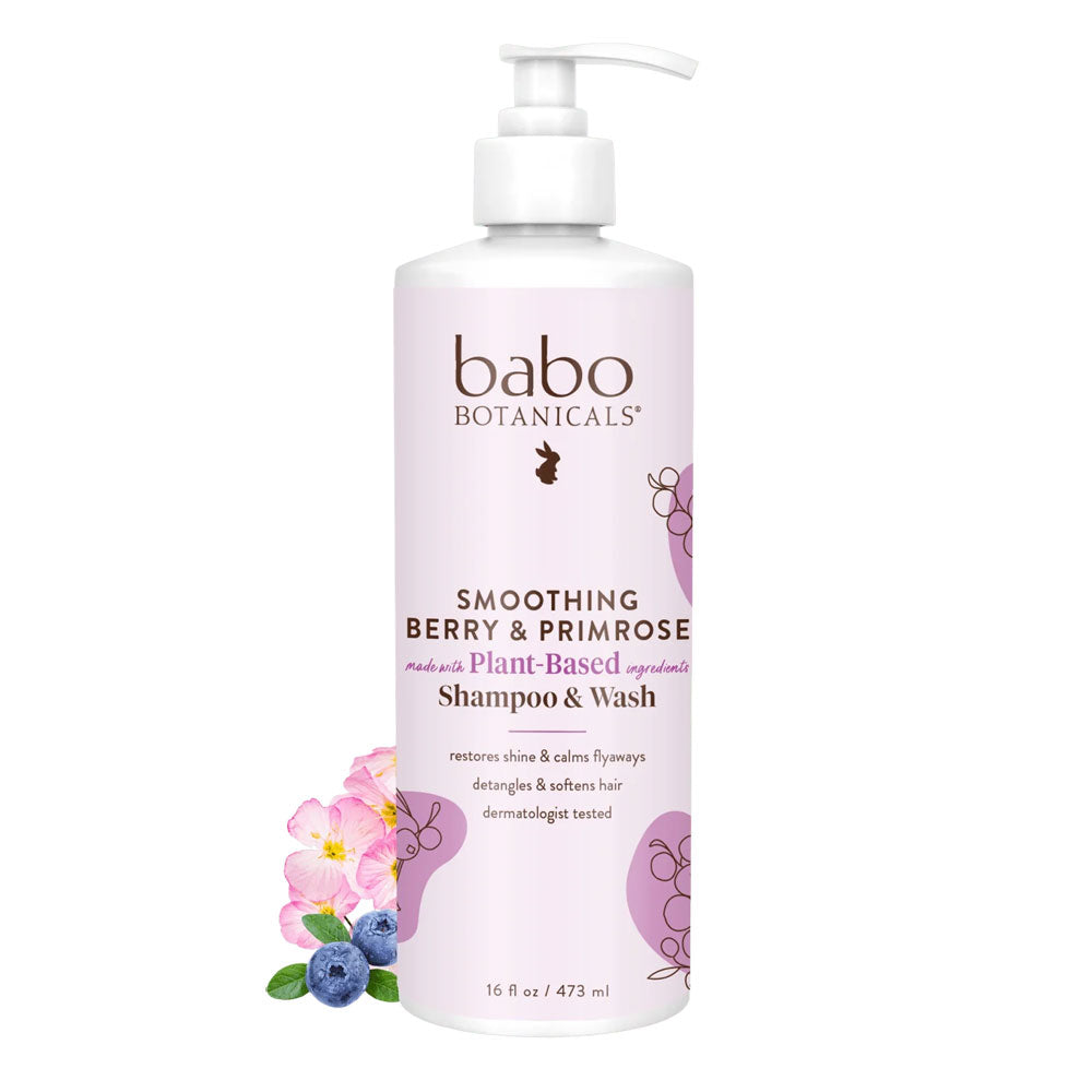 A pump bottle of Babo Botanicals Smoothing Berry & Primrose Shampoo & Wash with purple and green label design next to flowers and blueberries.