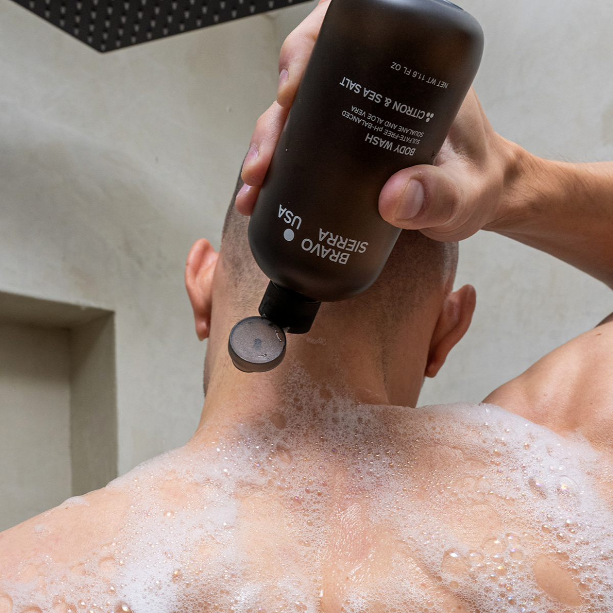 Man holding a bottle of citron & sea salt body wash over his soapy back in the shower. 