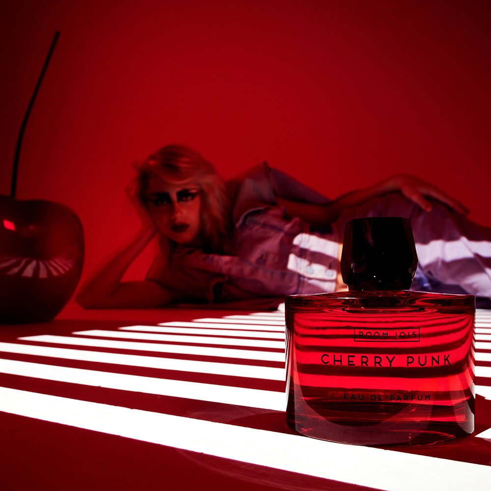Cherry Punk - Eau de Parfum bottle with a woman laying behind it with a large cherry prop and red background