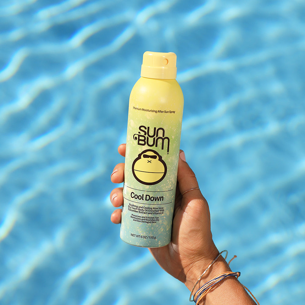 Sun Bum After Sun Cool Down Spray being held up against a backdrop of shimmering pool water.