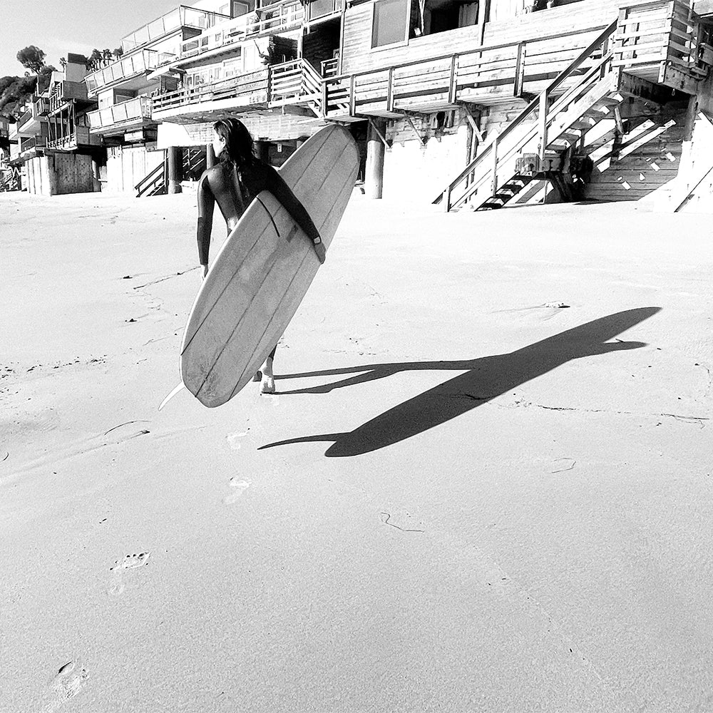 Woman in wetsuit with surfboard on a sandy beach facing wooden docks and staircase