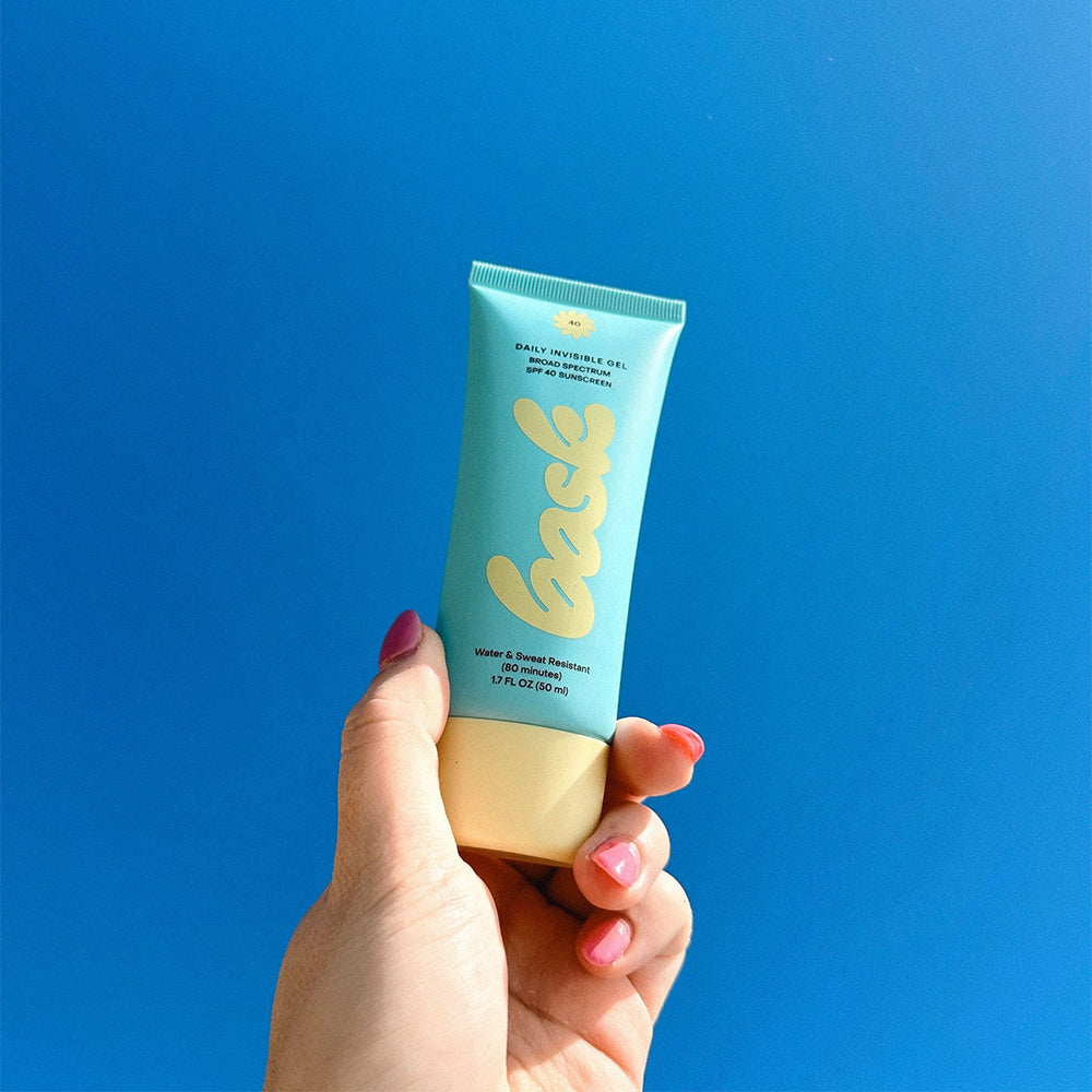 Hand holding a tube of Bask Daily Invisible Gel SPF 40 in front of a blue background
