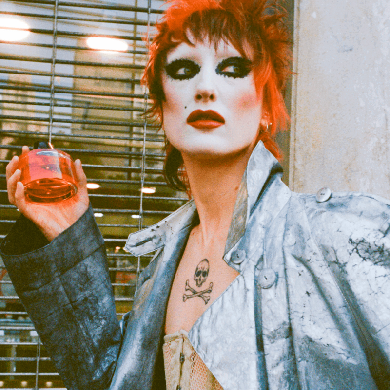 Punk rock person holding bottle of Room 1015 Jasmine Freak - Eau de Parfum in front of window blinds