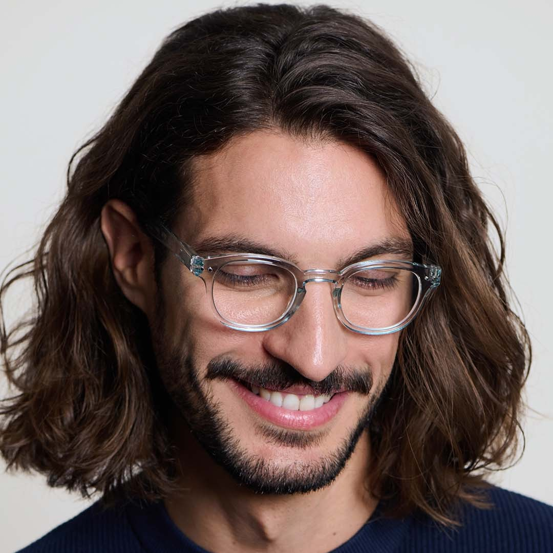 Man with long hair and glasses wearing a navy sweater against a white background